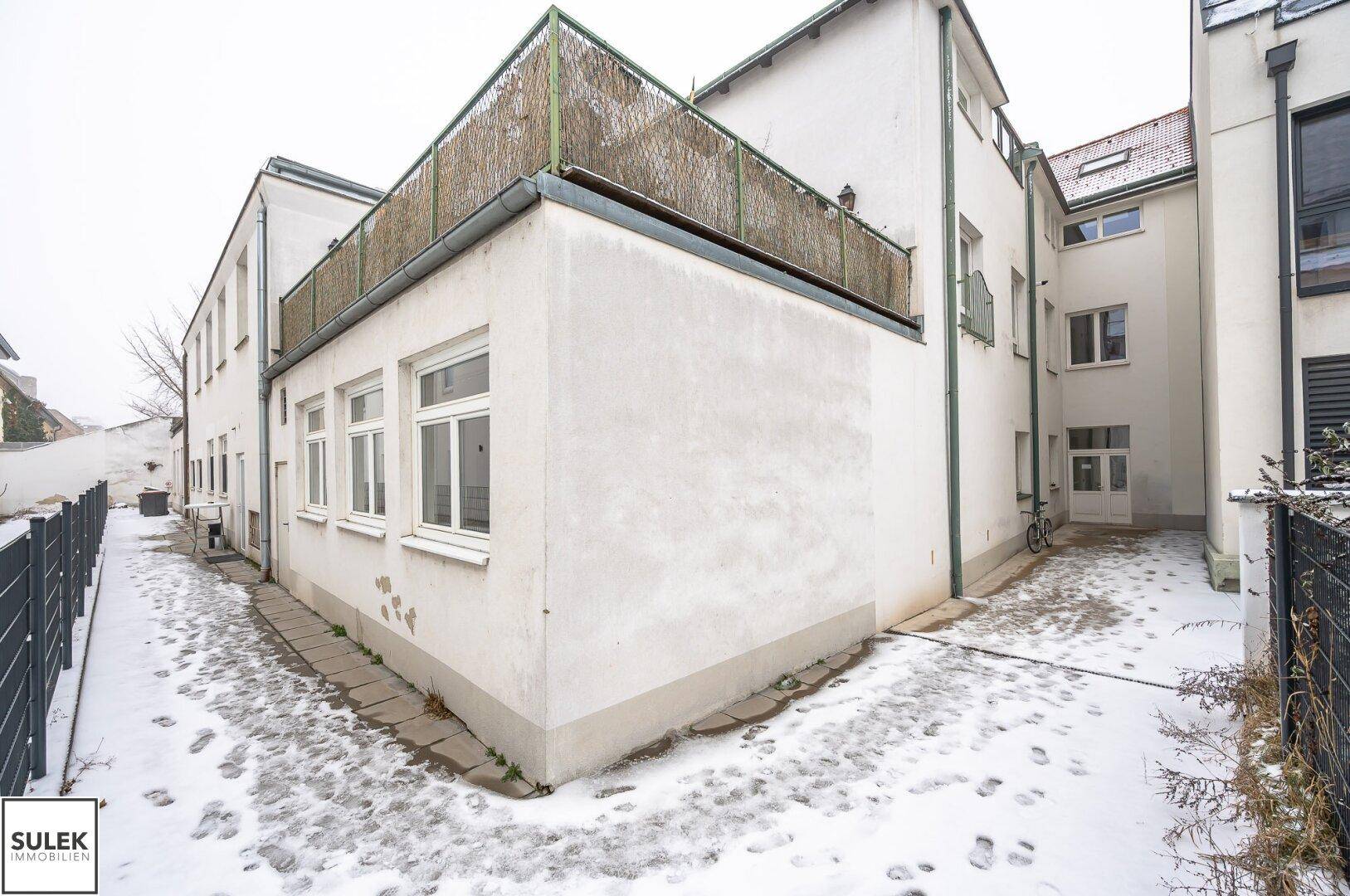 Hinterhof des Gebäudes mit verschneitem Boden und einer Terrasse im Obergeschoss mit Sichtschutz.