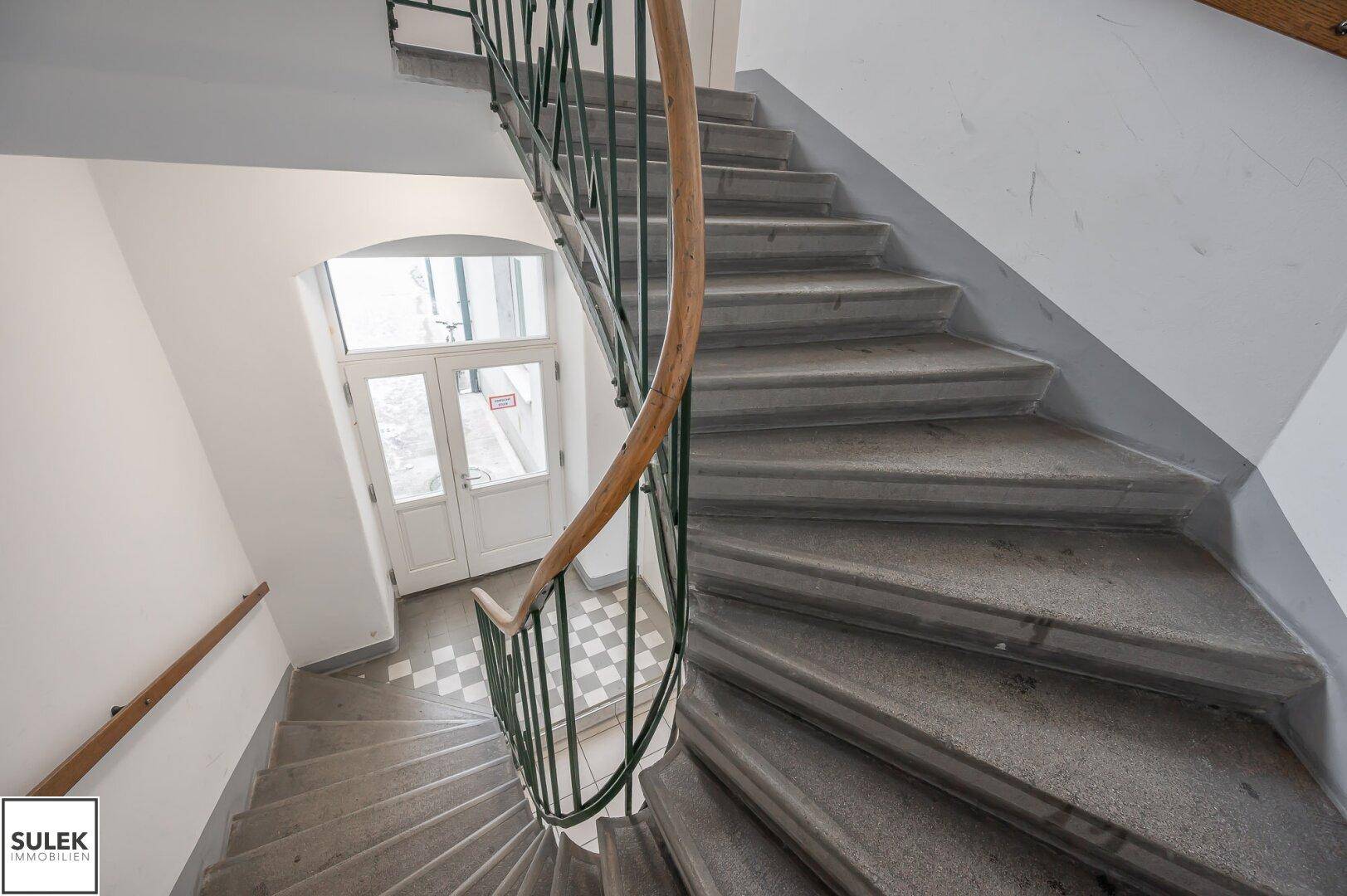 Wendeltreppe mit Steinstufen, grünem Metallgeländer und Holzhandlauf, führt zum Eingangsbereich.
