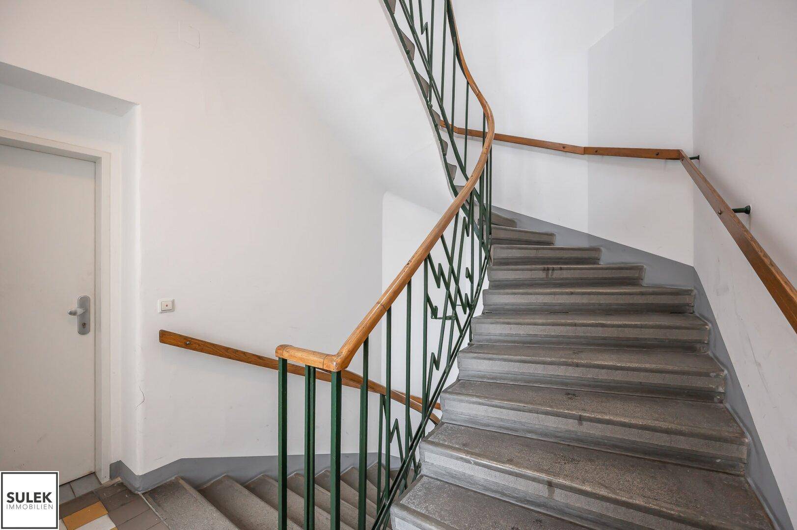 Treppenhaus mit Steinstufen, grünem Metallgeländer und Holzhandlauf in einem älteren Gebäude.