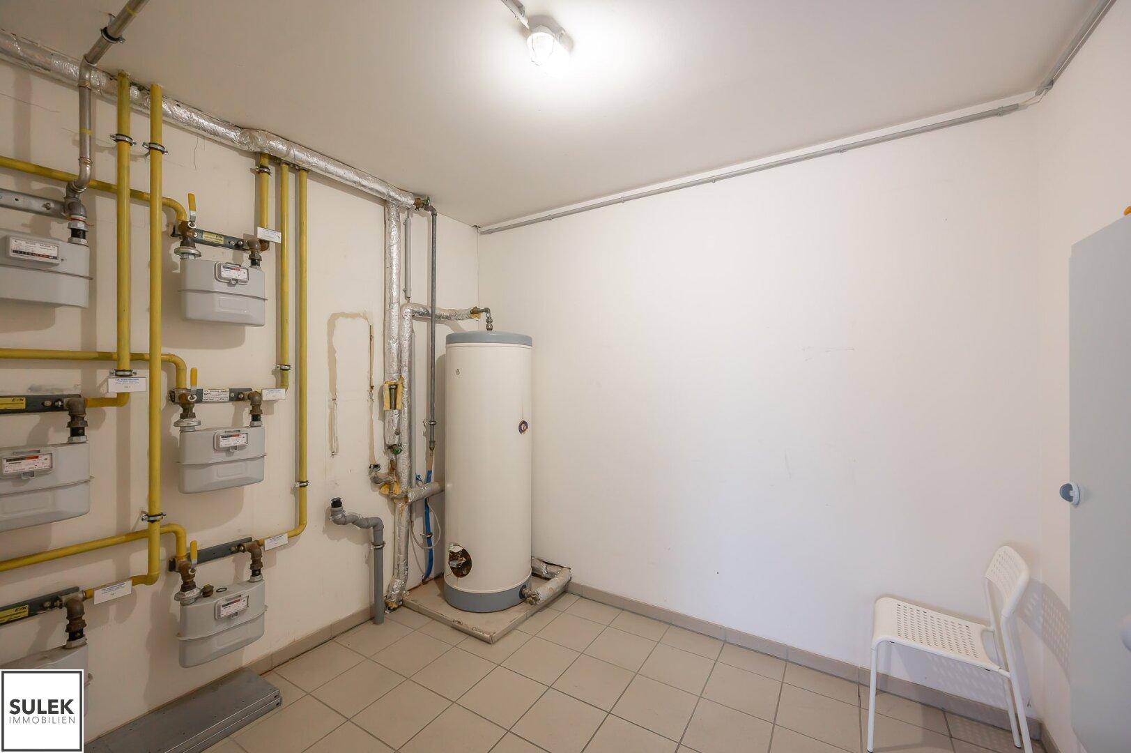 Technikraum mit Gaszählern, Warmwasserbereiter und Fliesenboden, funktional eingerichtet.