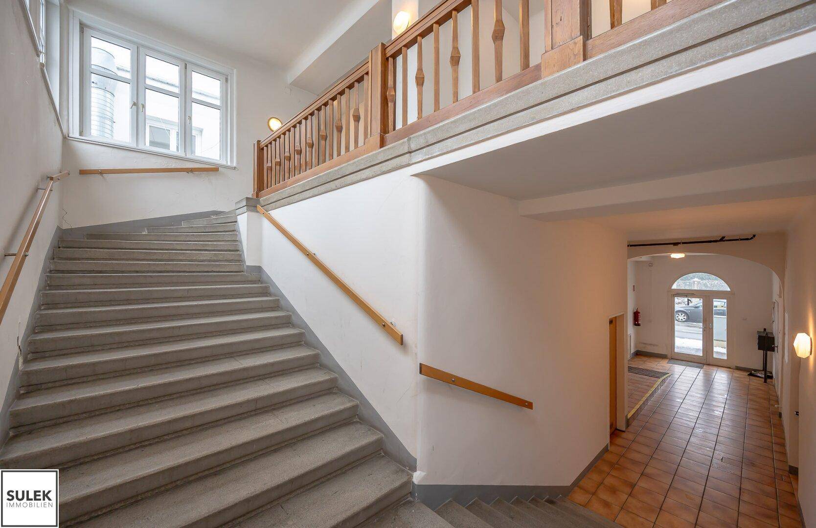 Breites Treppenhaus mit Steinstufen und Holzgeländer, führt zu einem gefliesten Flur.