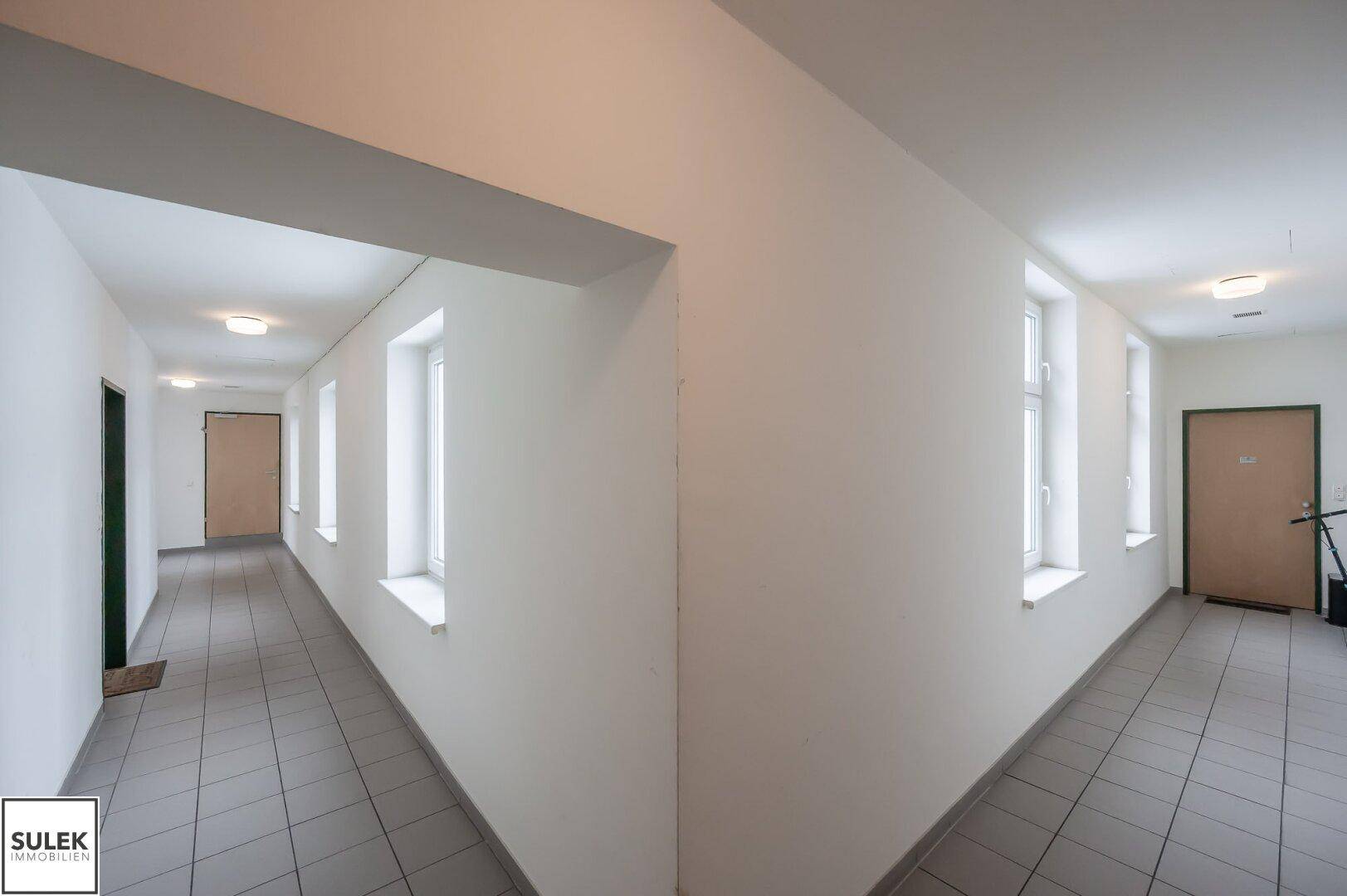 Heller Flur mit Fliesenboden und mehreren Türen, der sich um eine Ecke erstreckt.
