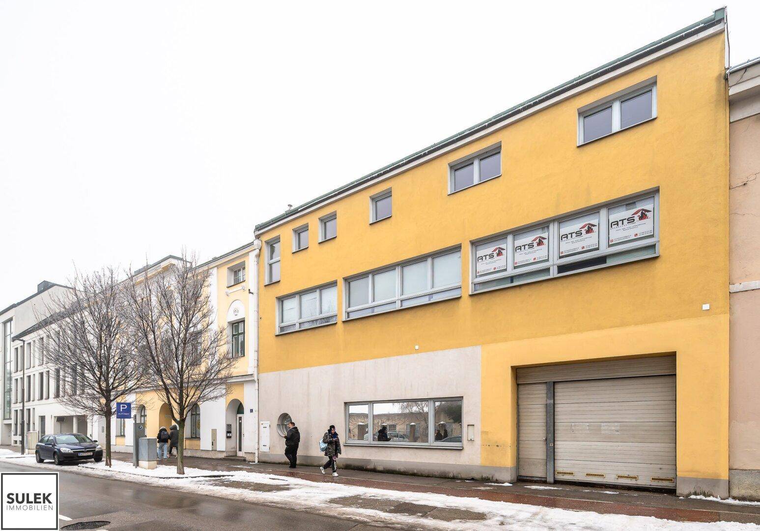 Gelbe Fassade eines Gebäudes mit großen Fenstern und einem Garagentor an der Straßenseite.