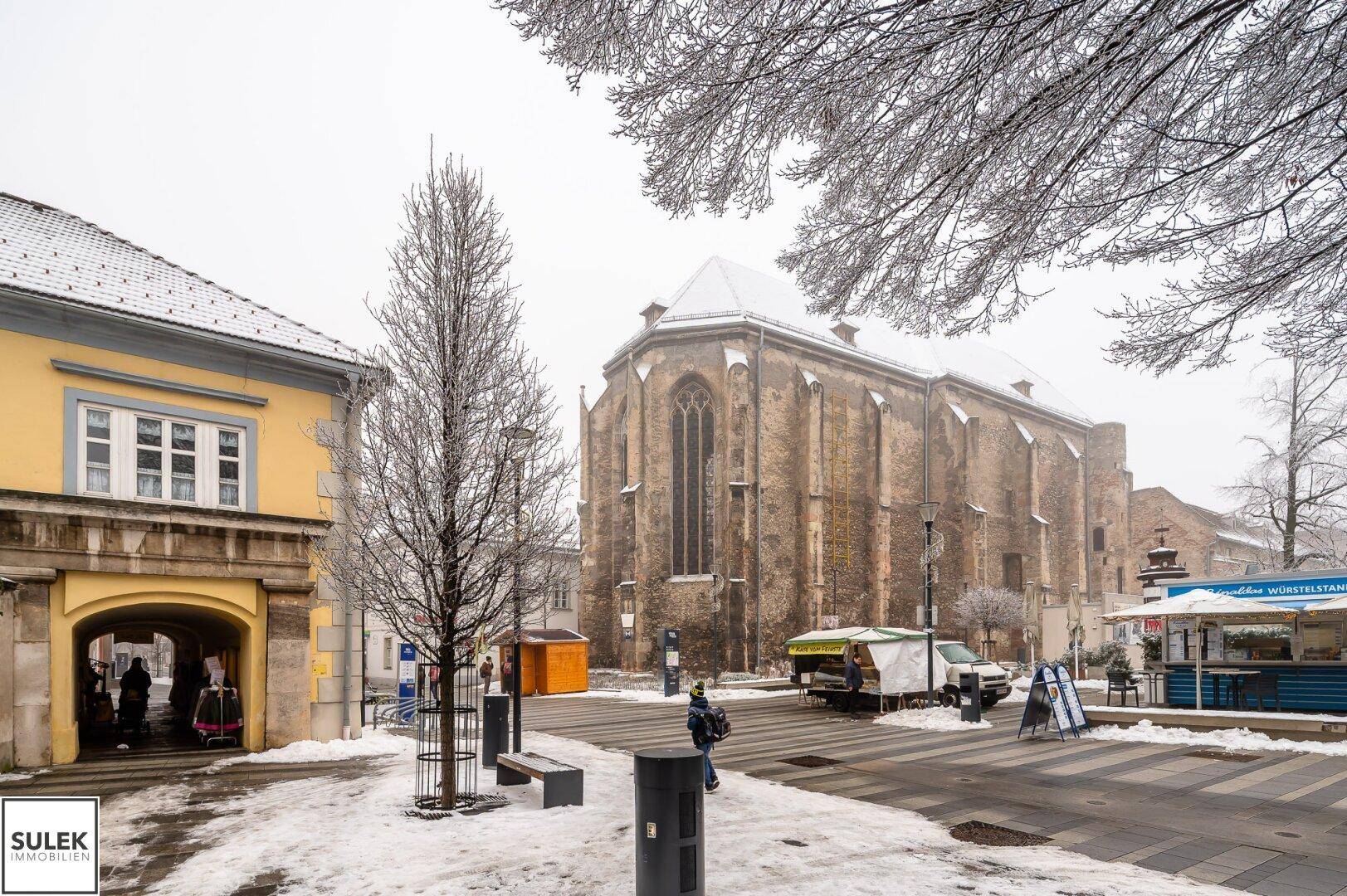 Winterliche Straßenszene mit einer historischen Kirche im Hintergrund und verschneiten Bäumen.