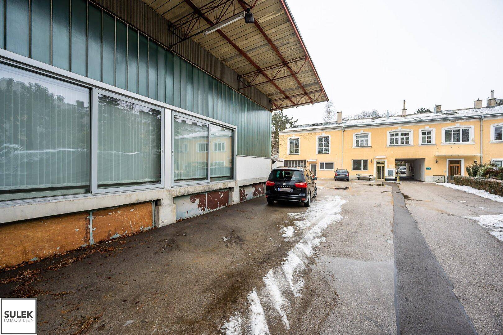 Ansicht der Gebäudefassade mit großen Fenstern und einem geparkten Auto, im Hintergrund ein weiteres gelbes Gebäude.