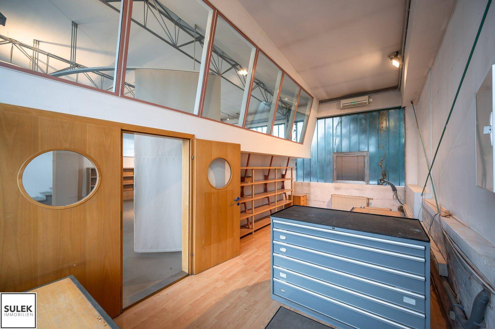 Heller Büroraum mit Holzboden, großen Fenstern und Regalen, ausgestattet mit einem blauen Aktenschrank.
