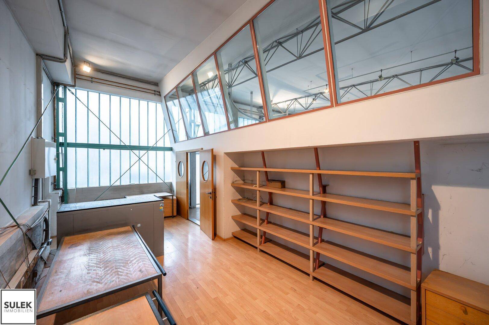 Geräumiger Bürobereich mit Holzboden, Regalen und großen Fenstern, die viel Tageslicht hereinlassen.