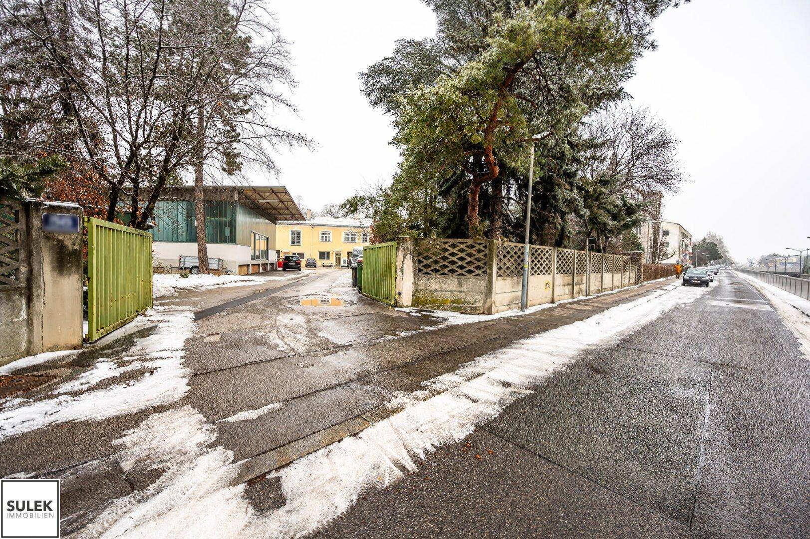 Zufahrt zum Grundstück mit offenem grünen Tor, umgeben von Bäumen und Schnee, mit Gebäuden im Hintergrund.