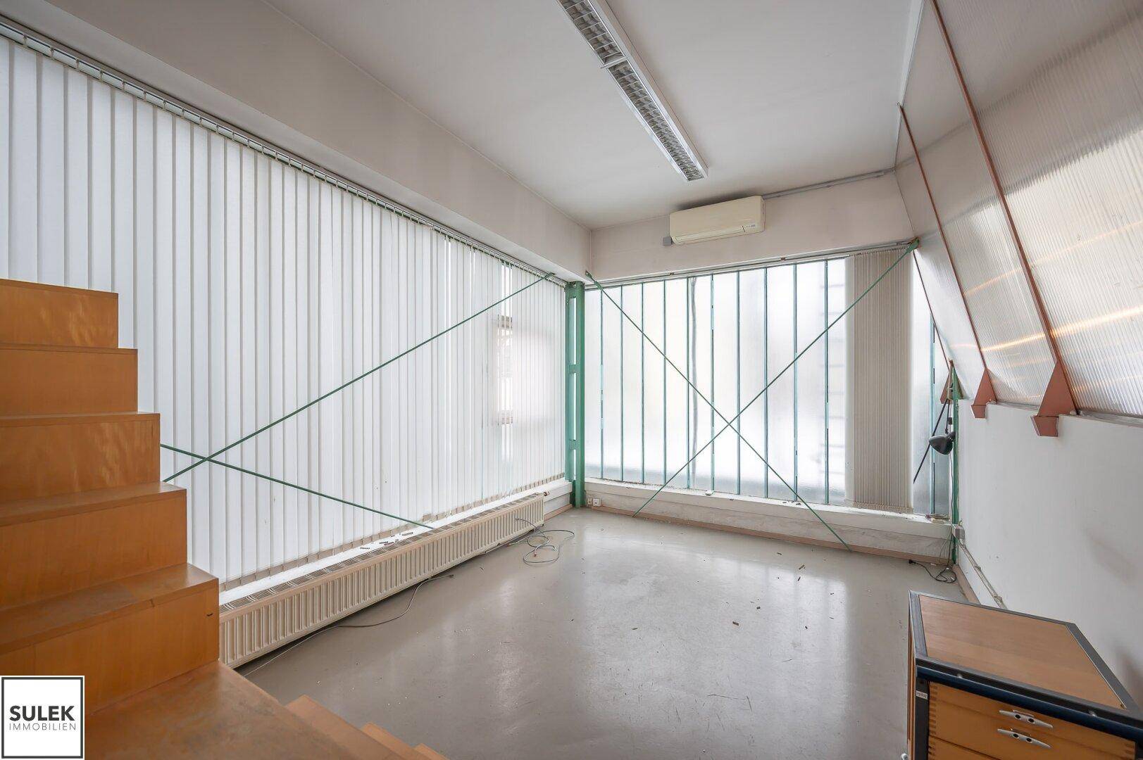 Heller Büroraum mit vertikalen Jalousien an großen Fenstern und einem Betonboden, ausgestattet mit Heizkörpern.