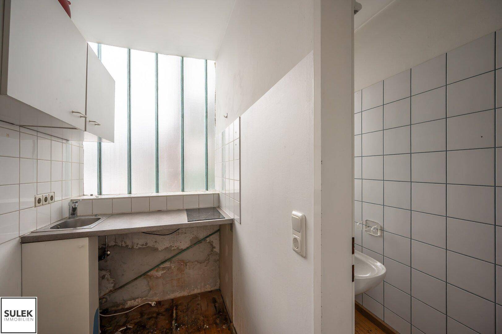 Kleine Küchenzeile mit Spüle und Kochfeld, angrenzend an einen Toilettenbereich mit Fliesen.