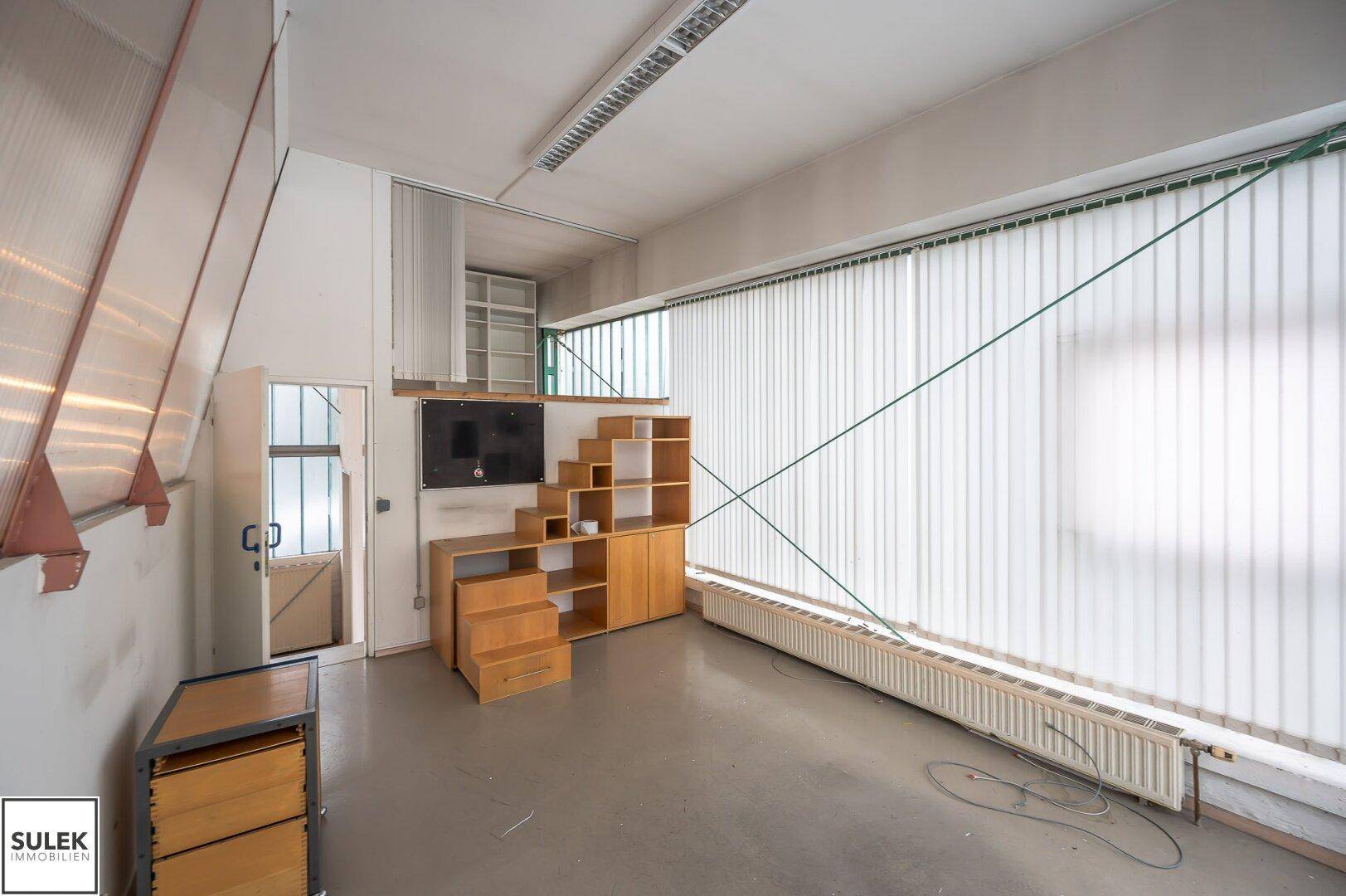 Büroraum mit großen Fenstern, vertikalen Jalousien und einem Betonboden, ausgestattet mit Holzregalen und einem Fernseher.