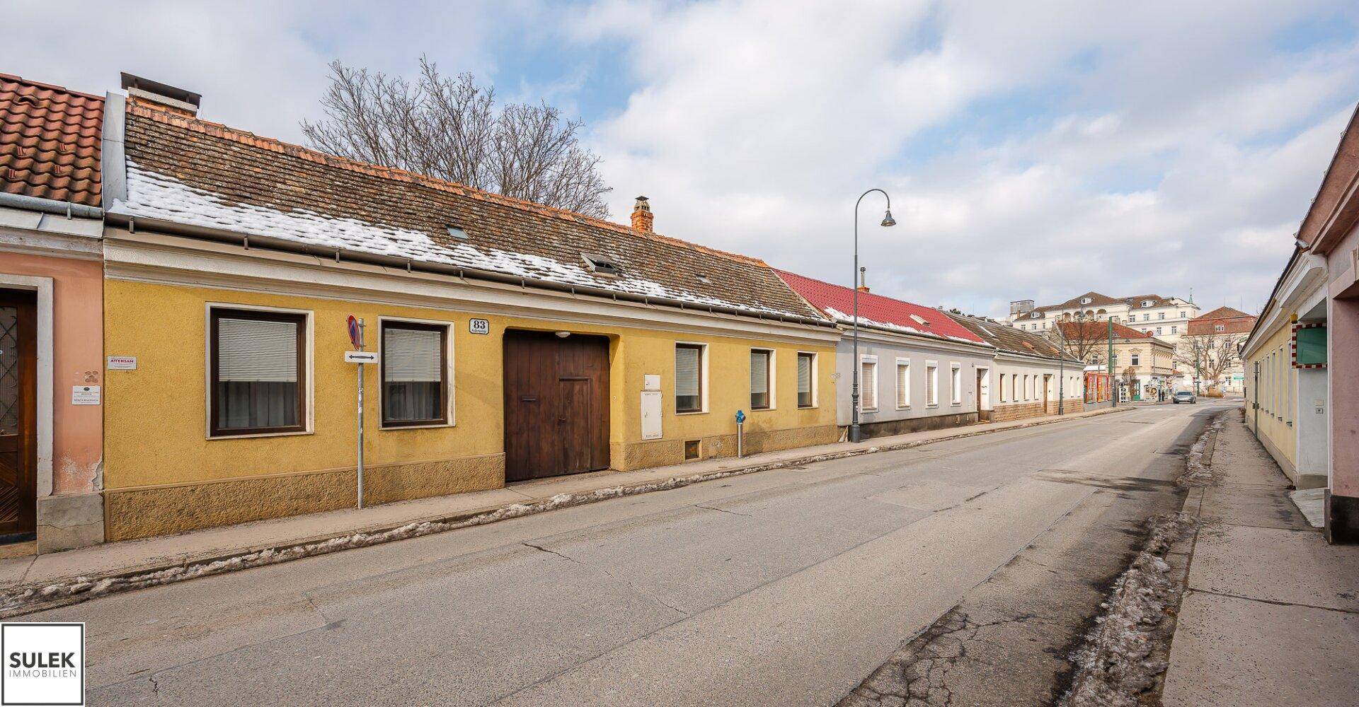 Blick auf die Straße mit der gelben Hausfassade und angrenzenden Gebäuden unter bewölktem Himmel.