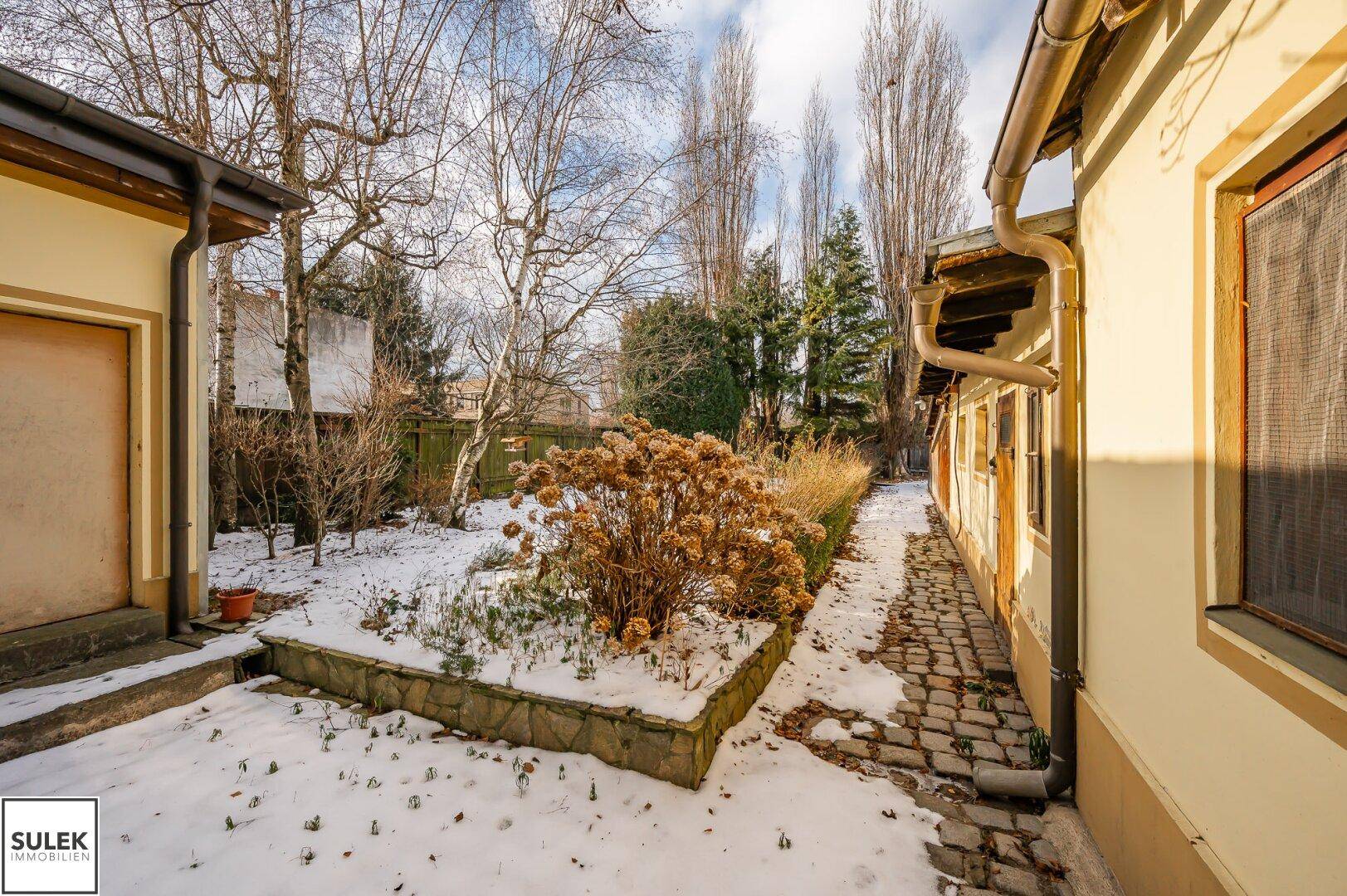 Verschneiter Gartenbereich mit Beeten, Sträuchern und Blick auf die umliegenden Gebäude.