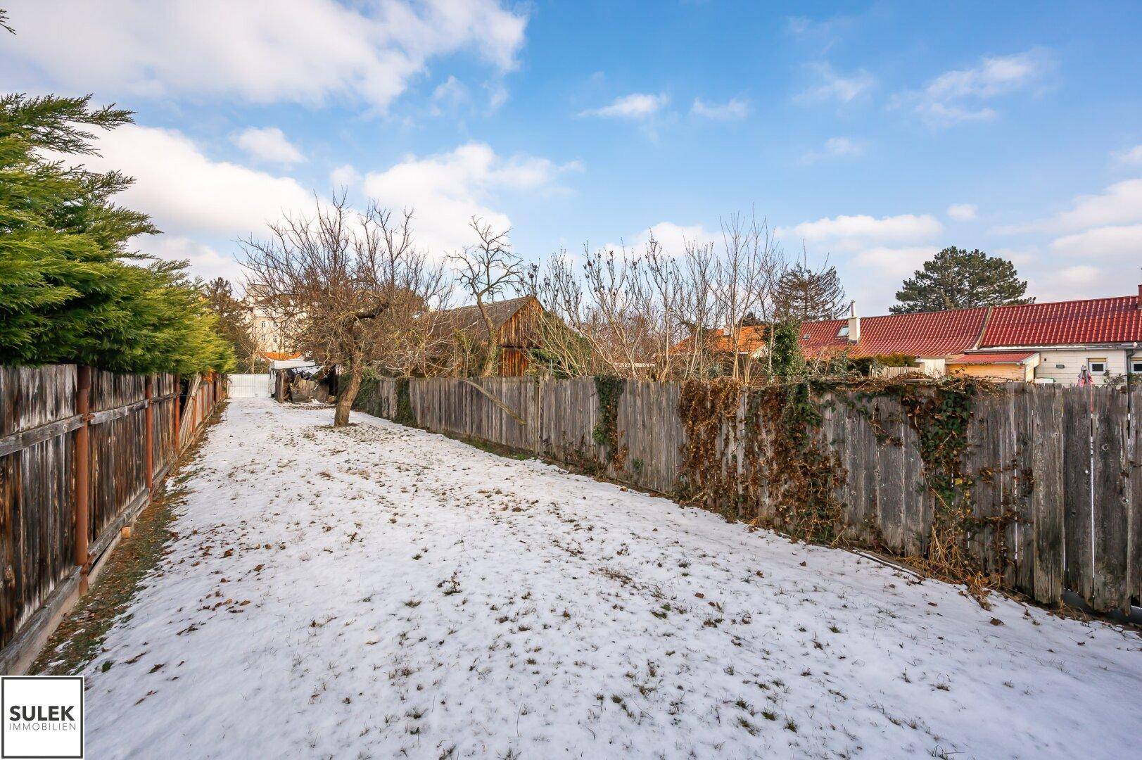 Weitläufiger, verschneiter Gartenbereich mit Holzzaun und Blick auf weitere Gebäude im Hintergrund.