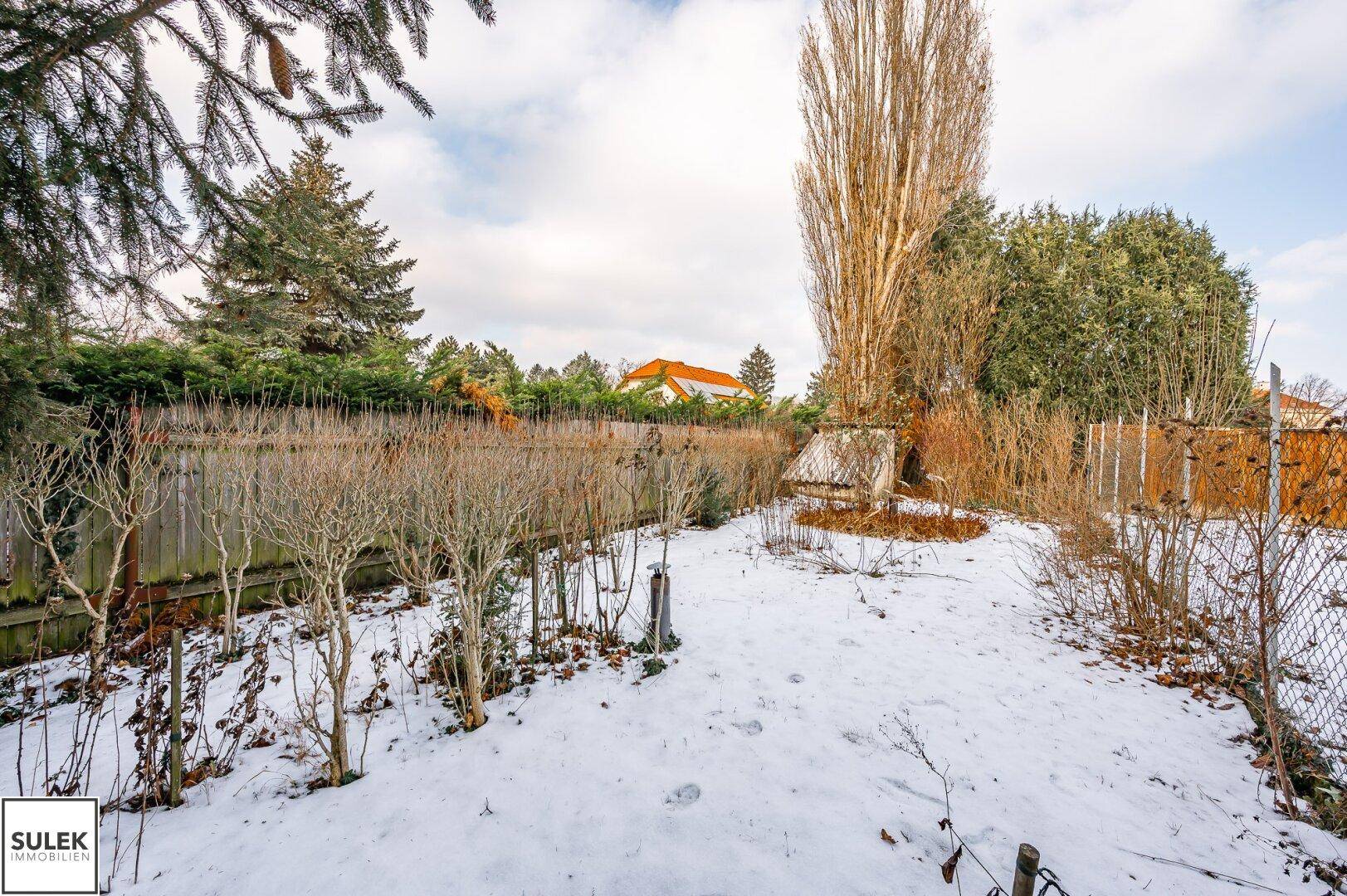 Verschneiter Gartenbereich mit Sträuchern und Bäumen, umgeben von einem Holzzaun.