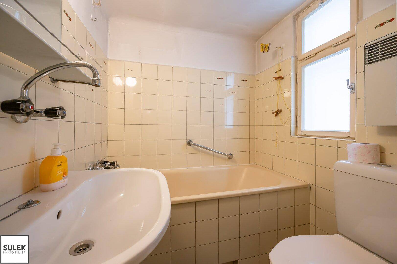 Badezimmer mit Badewanne, WC und Fenster, ausgestattet mit hellen Fliesen und Haltegriffen.