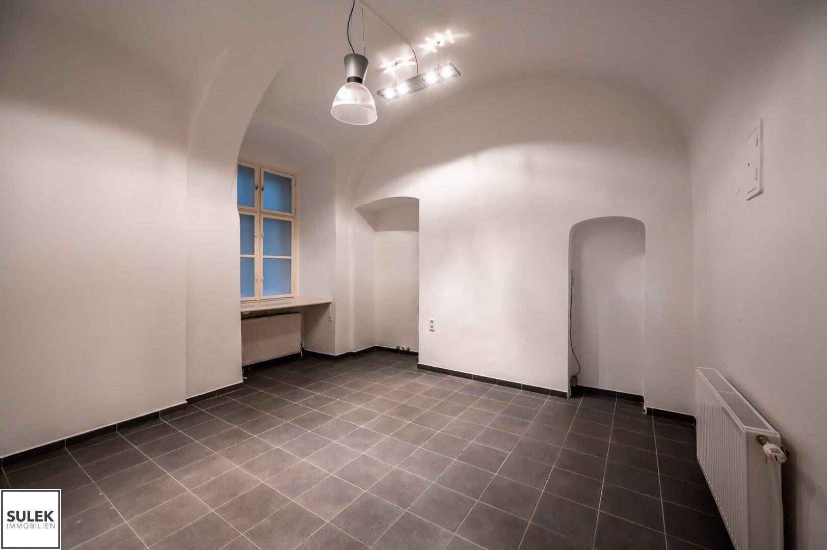 Weiterer heller Raum mit gewölbter Decke, Fenster und dunklen Bodenfliesen.
