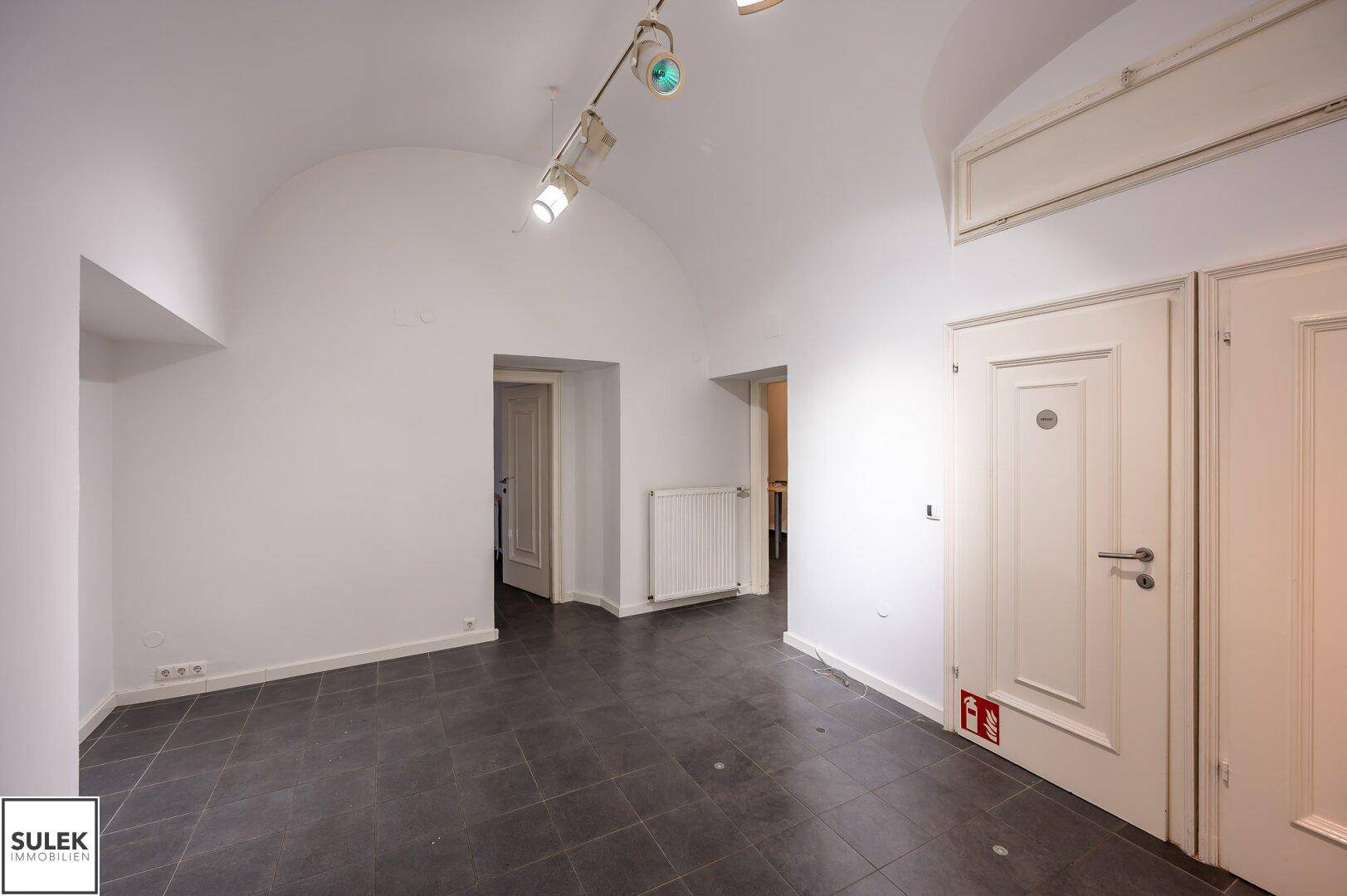 Heller Geschäftsraum mit gewölbter Decke, dunklen Fliesen und Zugang zu weiteren Räumen.