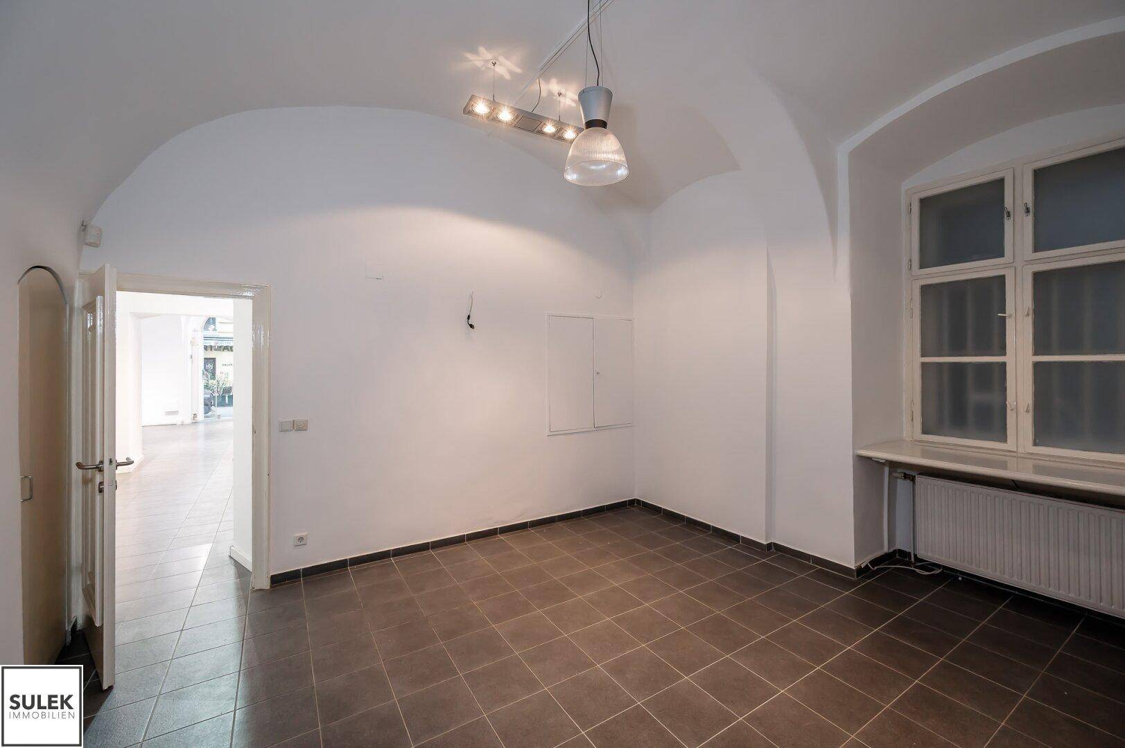 Geräumiger Raum mit gewölbter Decke, Fenster und dunklen Bodenfliesen.