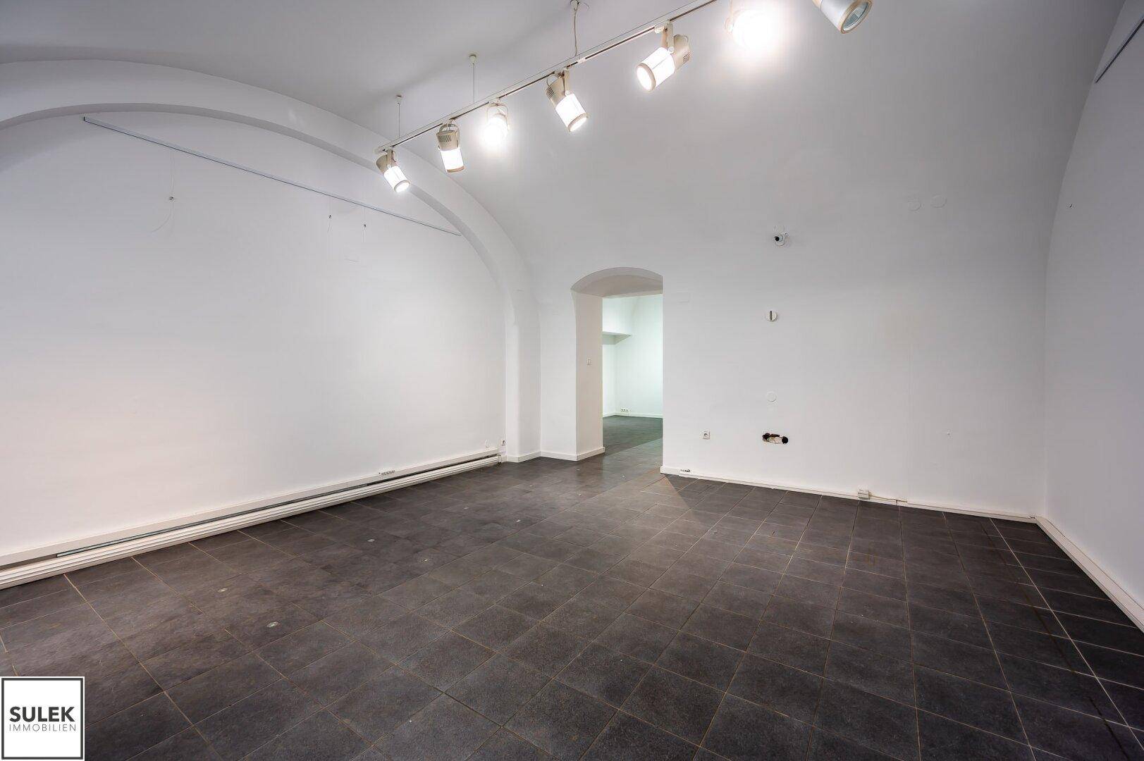 Langer, heller Geschäftsraum mit gewölbter Decke und dunklen Bodenfliesen.