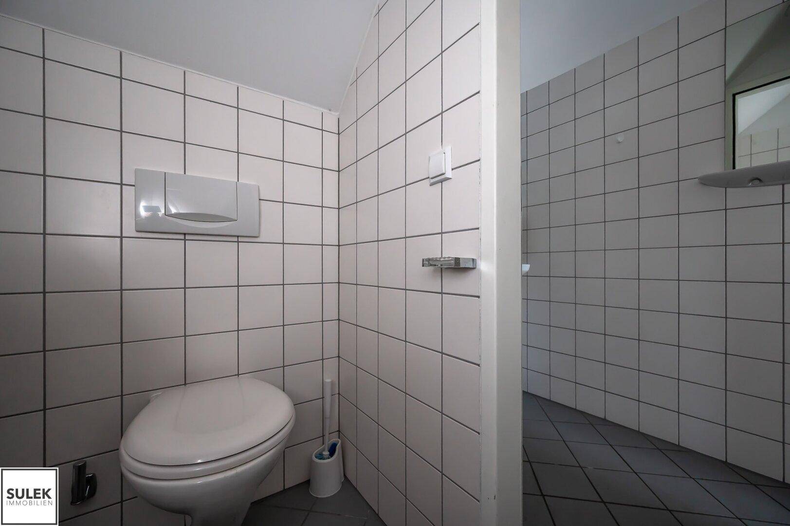 Einfache Toilette mit weißen Fliesen an den Wänden und grauem Fliesenboden.