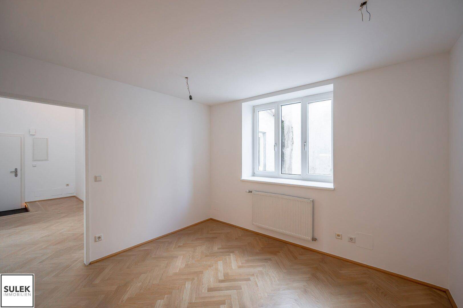 Helles Zimmer mit Fischgrätparkett und einem Fenster, das den Raum mit Tageslicht versorgt.