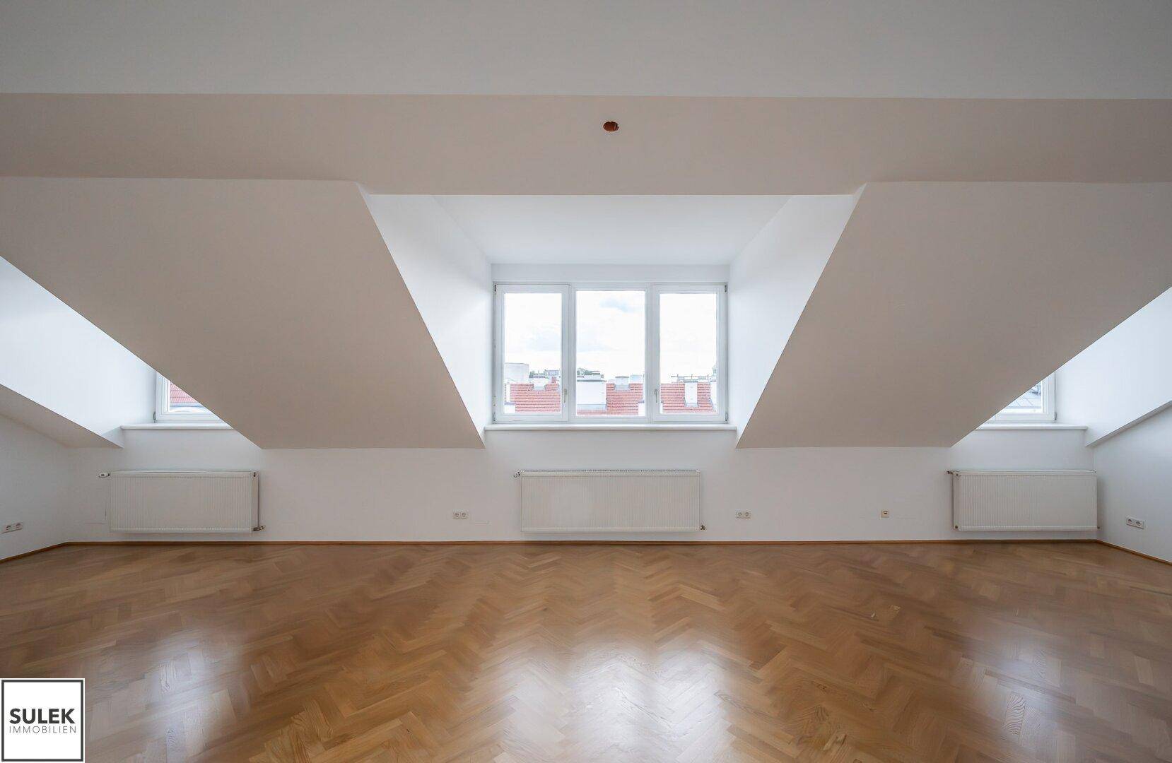 Heller Wohnraum im Dachgeschoss mit Fischgrätparkett und großen Fenstern, die viel Tageslicht hereinlassen.