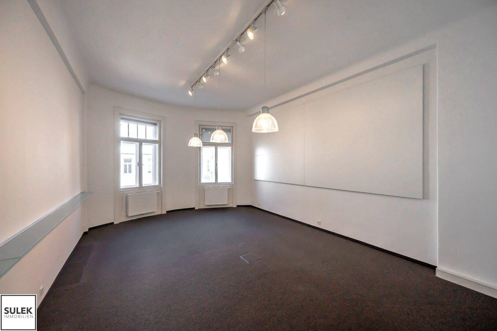 Kleinerer Büroraum mit Teppichboden, Deckenstrahlern und zwei Fenstern, die Tageslicht spenden.