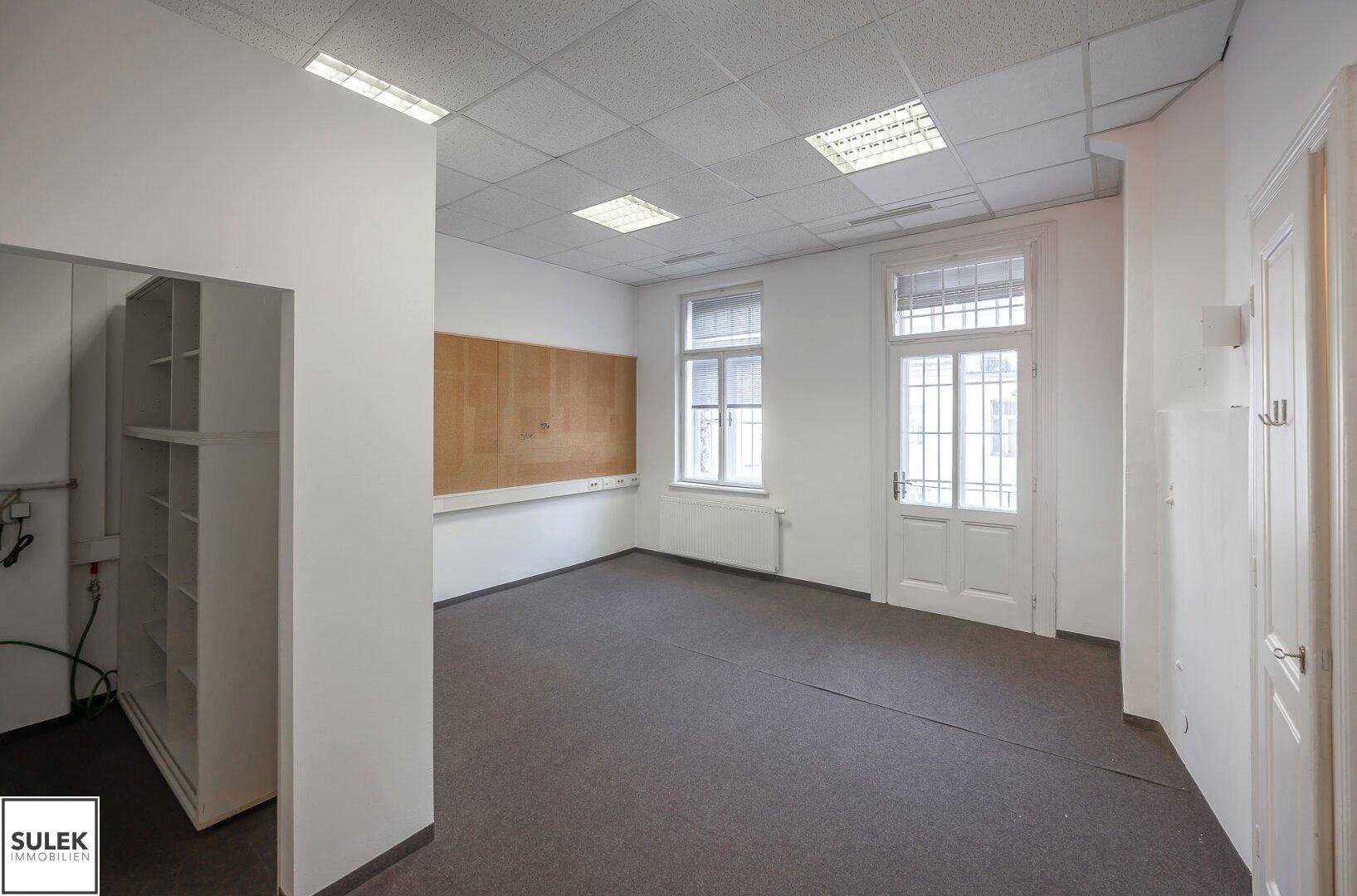 Büroraum mit Teppichboden, Regalen und einer großen Korktafel an der Wand.