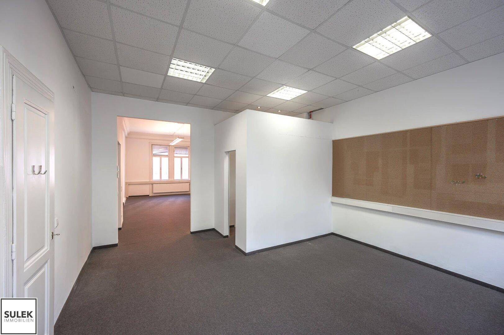 Büroraum mit abgehängter Decke, Teppichboden und einer großen Korktafel an der Wand.
