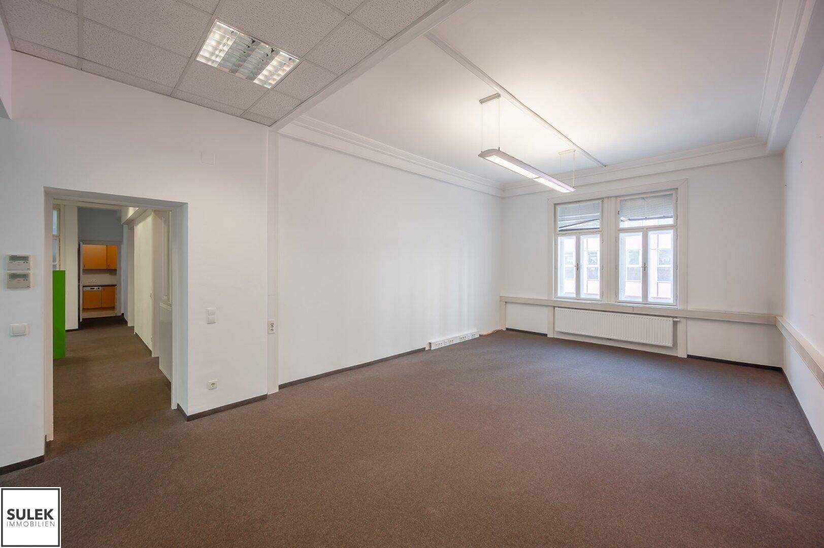 Geräumiger Büroraum mit Teppichboden, abgehängter Decke und großen Fenstern.