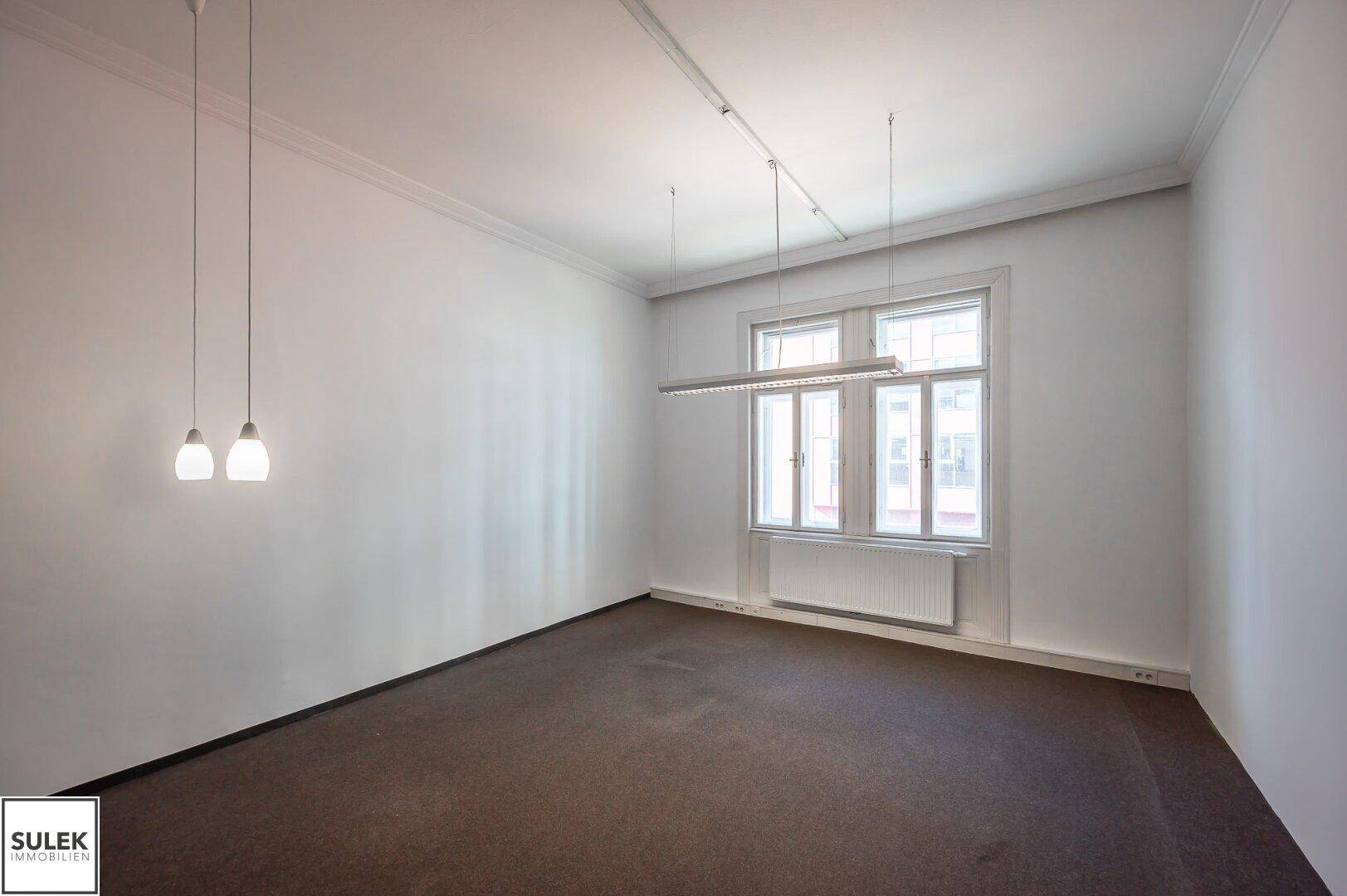 Heller Büroraum mit Teppichboden, modernen Hängelampen und großen Fenstern.