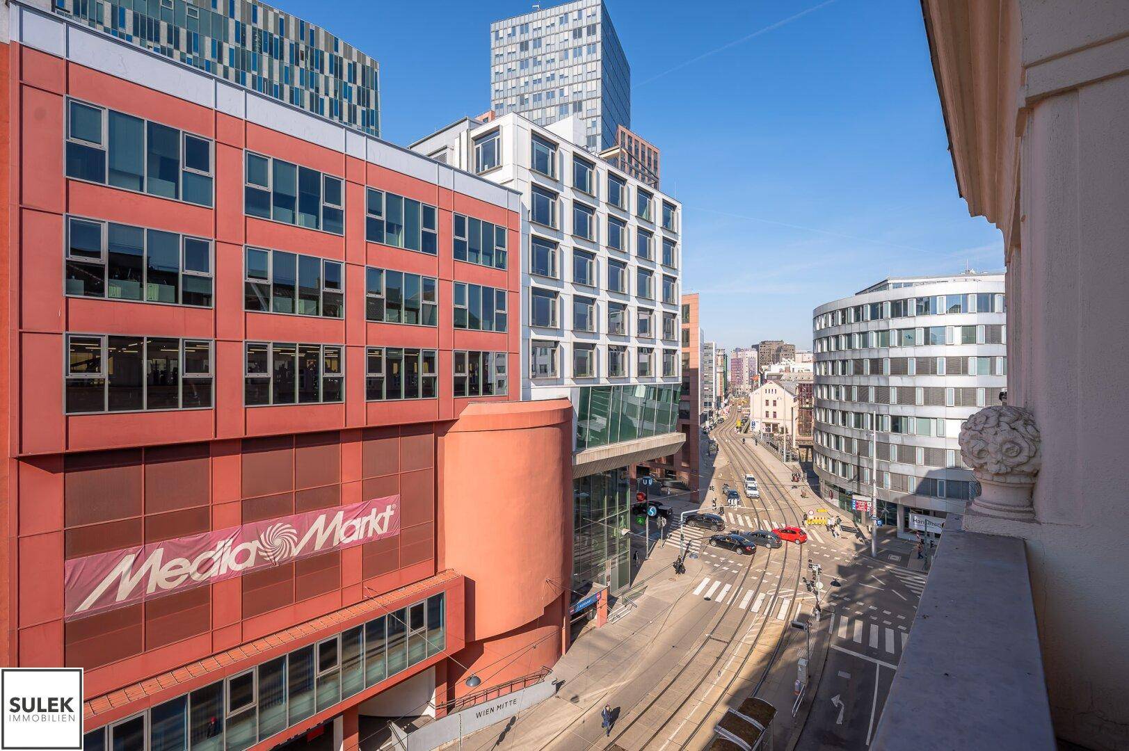 Blick auf eine belebte Stadtstraße mit modernen Gebäuden und Straßenbahnschienen.