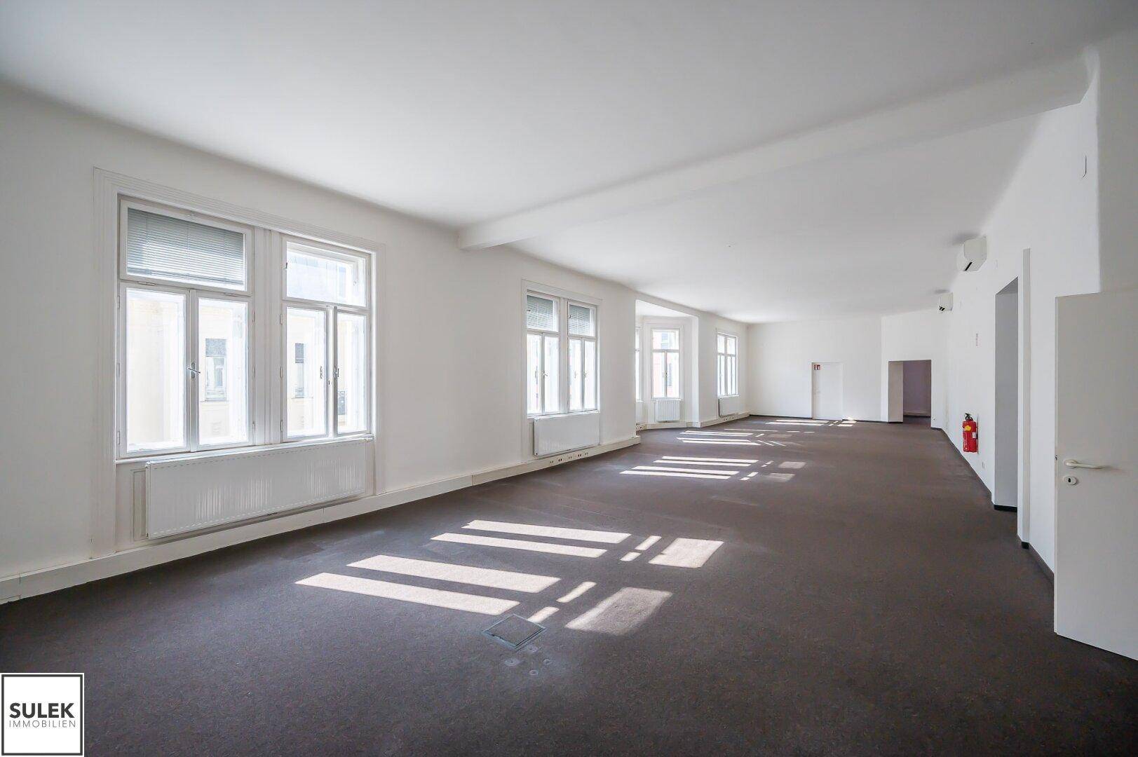 Heller und weitläufiger Büroraum mit Teppichboden und mehreren Fenstern, die den Raum gut belichten.