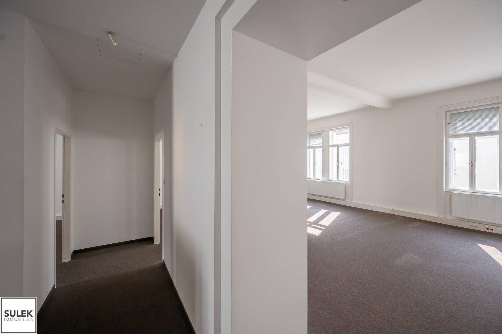 Heller Flur mit Teppichboden, der zu einem großen Bürobereich mit Fenstern führt.