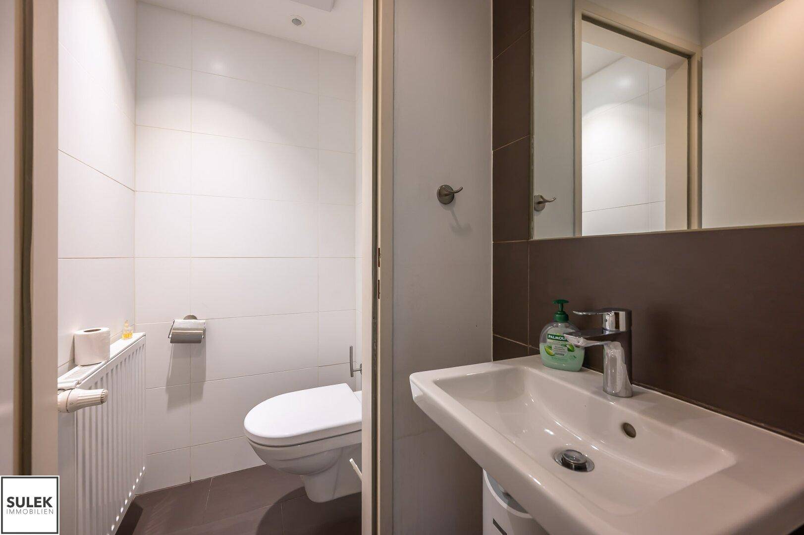 Helles Badezimmer mit weiß gefliesten Wänden, einem Waschbecken und einer Toilette.