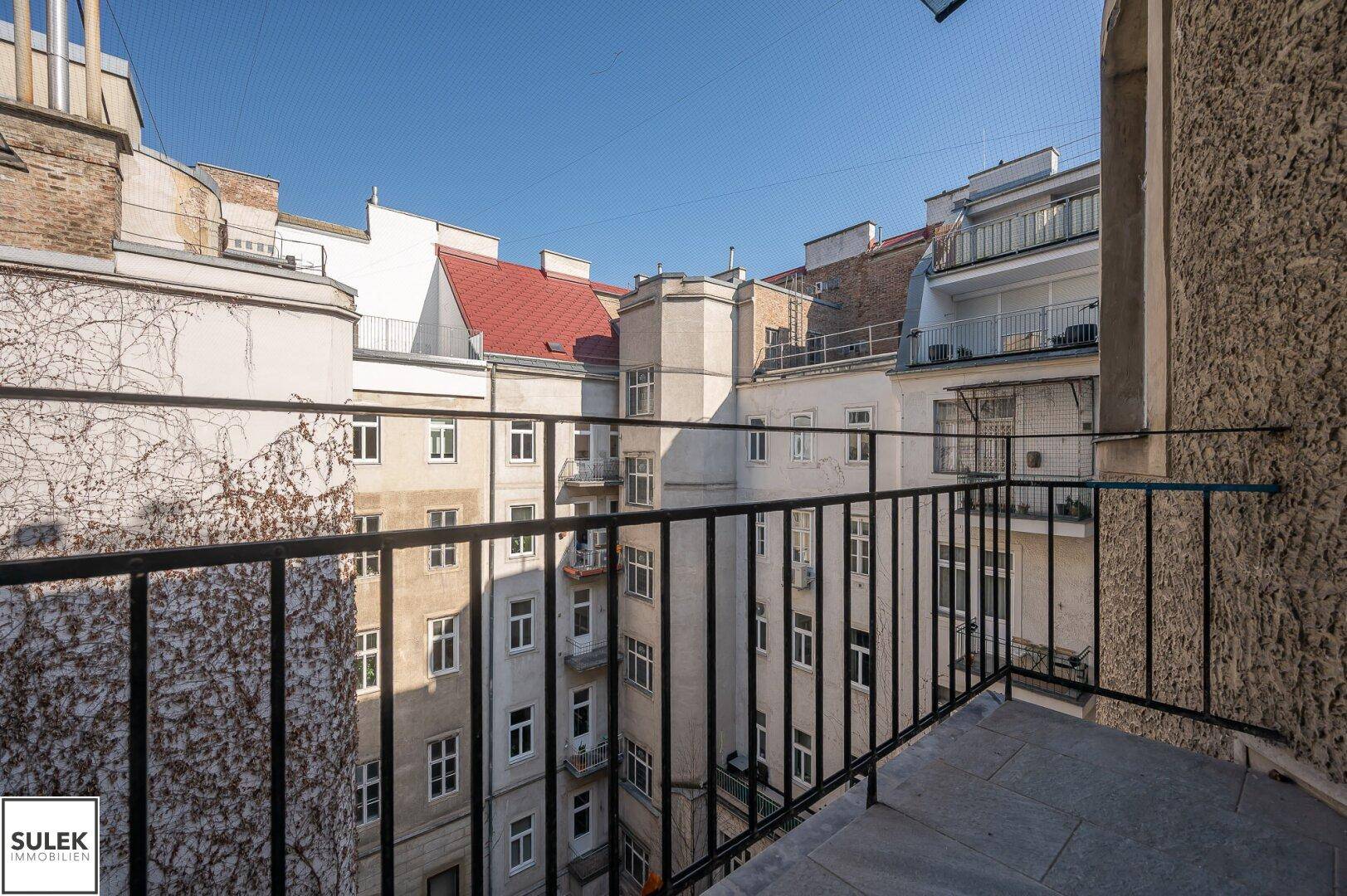 Kleiner Balkon mit Metallgeländer und Blick auf den Innenhof eines Altbaus.