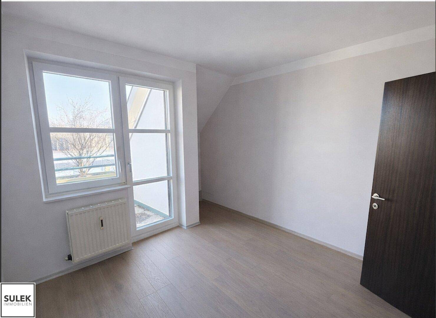 Helles Schlafzimmer mit Laminatboden und großem Fenster, das viel Tageslicht hereinlässt.