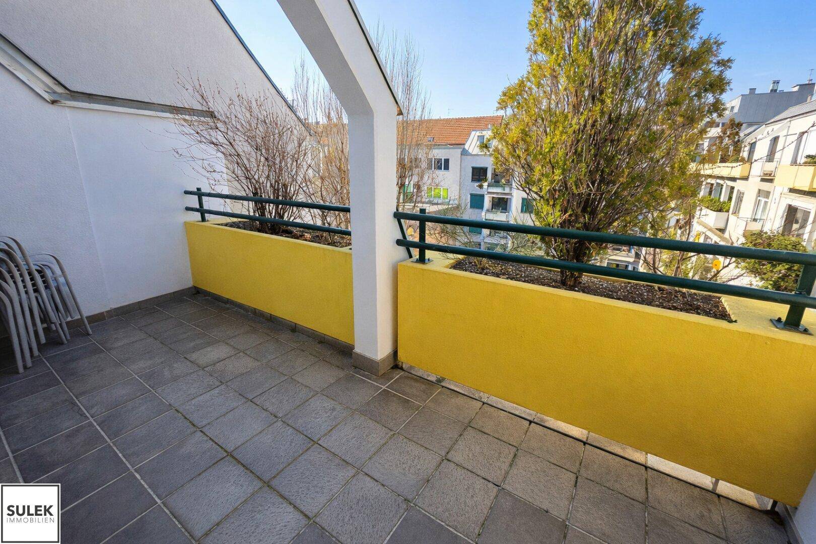 Sonniger Balkon mit Fliesenboden, gelben Pflanzkästen und Blick auf die Umgebung.