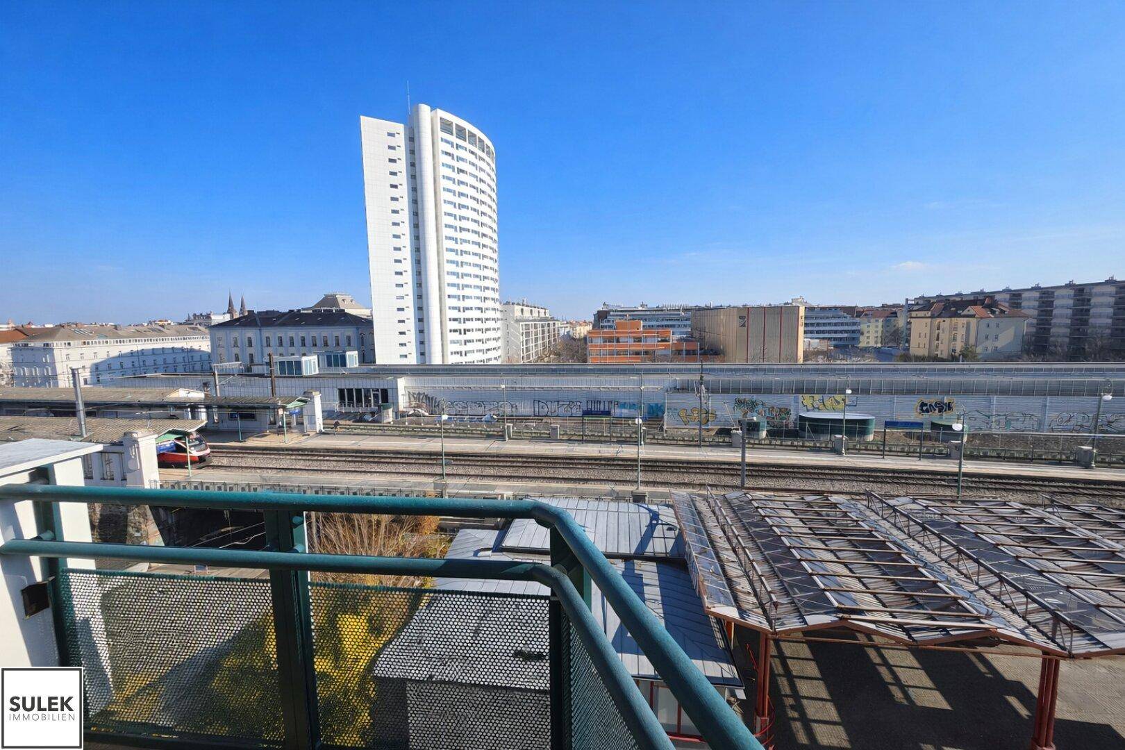 Weitläufiger Blick über Bahngleise und die Stadt mit einem markanten Hochhaus im Hintergrund.