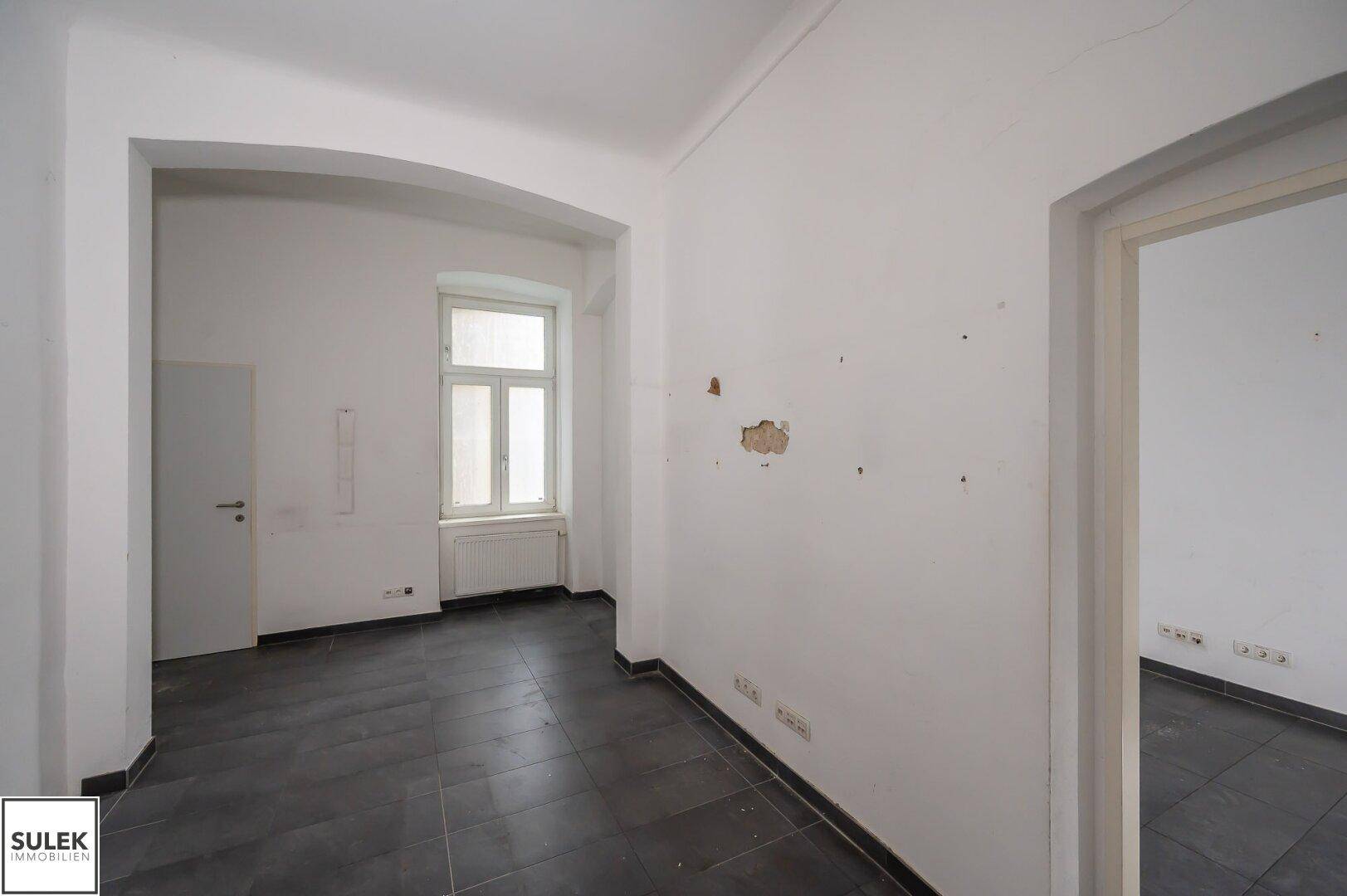 Heller Büroraum mit dunklem Fliesenboden, weißen Wänden und einem Fenster, das Tageslicht spendet.