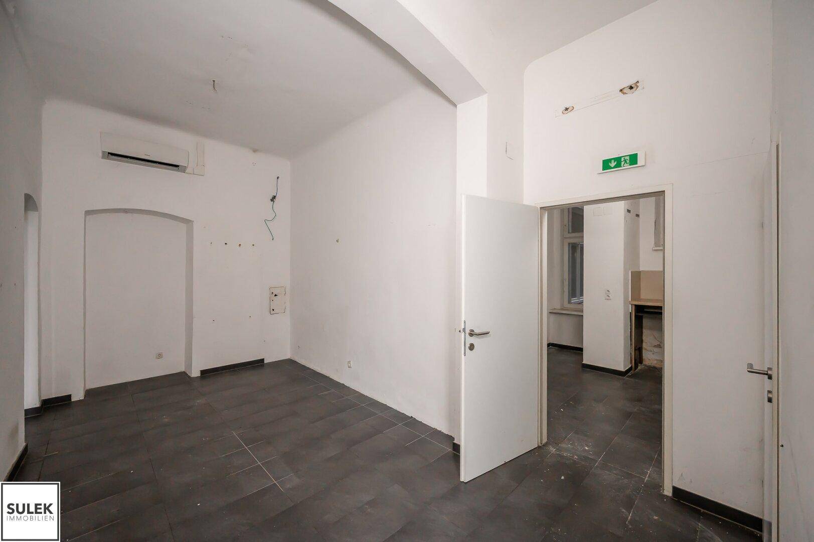 Leerer Büroraum mit dunklen Fliesen, weißen Wänden und einer Klimaanlage an der Decke.