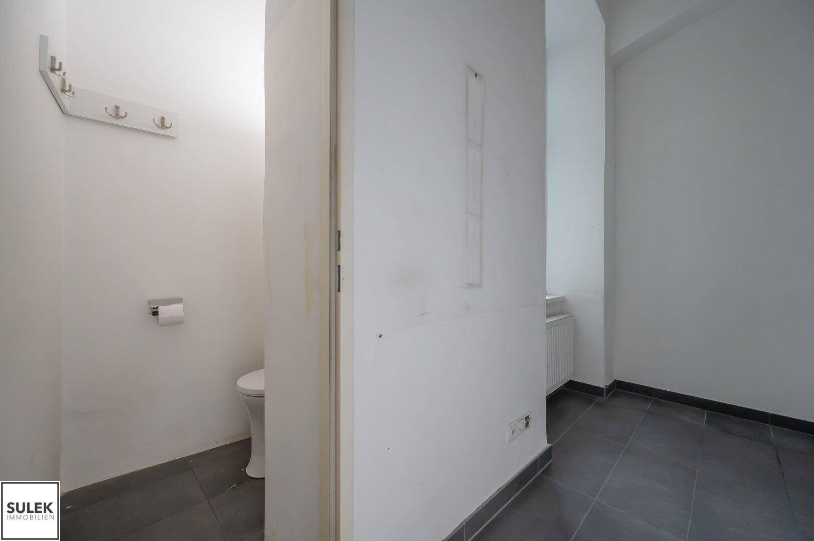 Einfache Toilette mit weißer Keramik und einem Kleiderhaken an der Wand, in einem kleinen Raum.