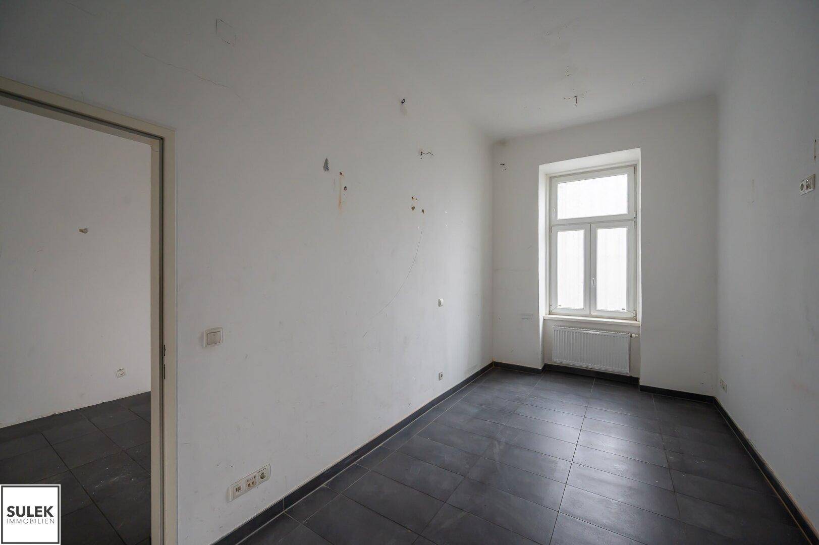 Leerer Büroraum mit dunklen Fliesen, weißen Wänden und einem Fenster mit Heizkörper darunter.