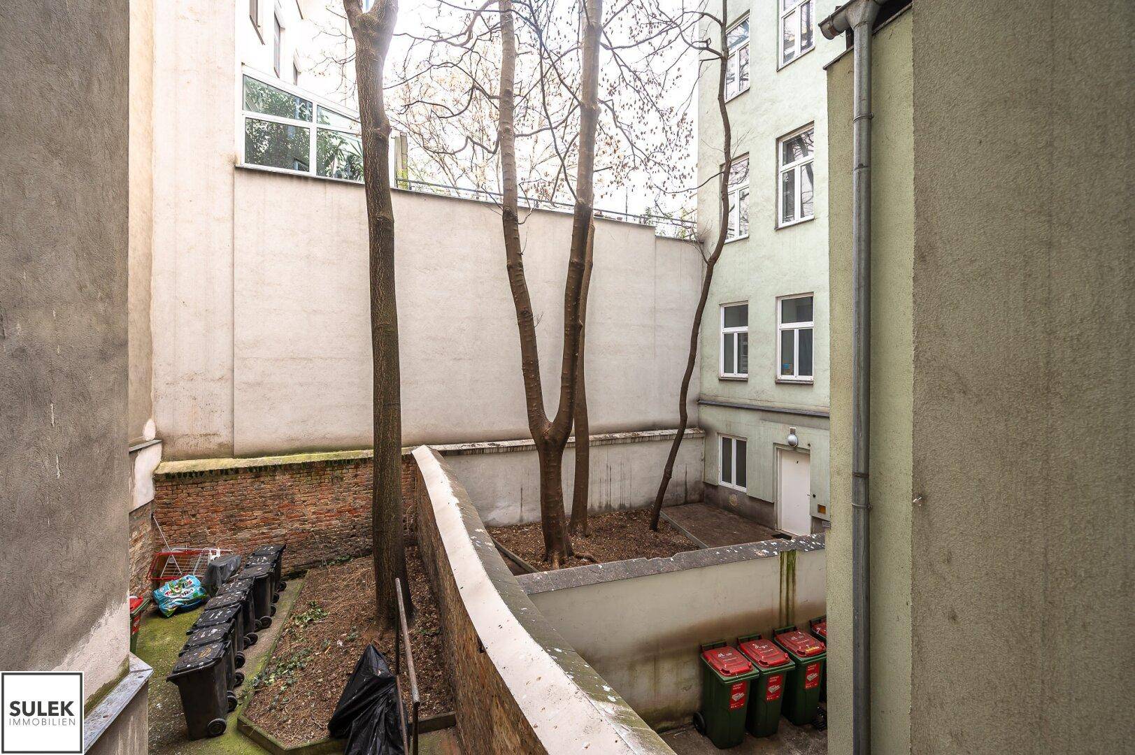 Innenhof mit Bäumen, Mülltonnen und hohen Mauern, der einen Blick auf die Rückseite des Gebäudes bietet.