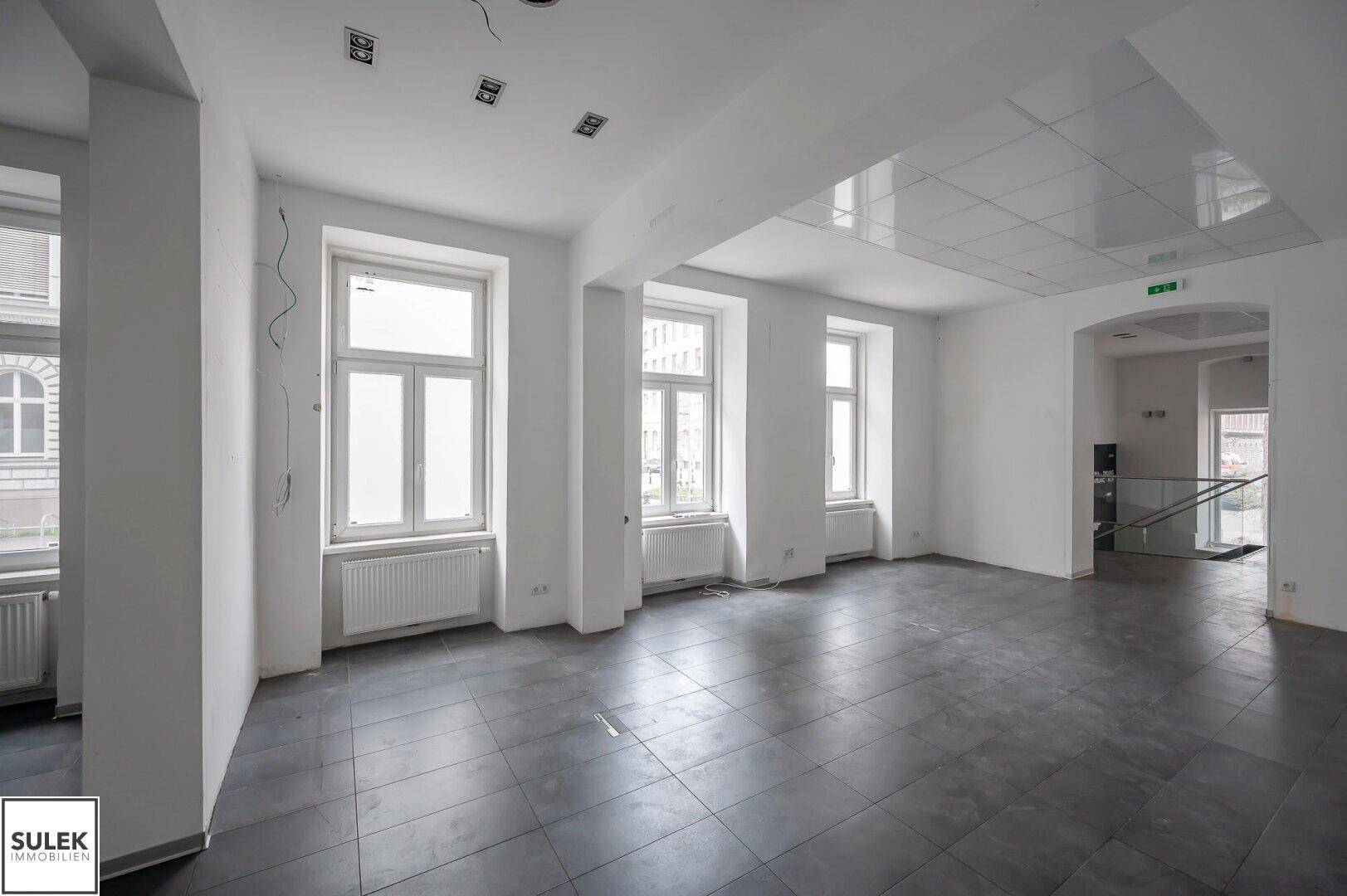Geräumiger Büroraum mit großen Fenstern, dunklen Fliesen und einer abgehängten Decke.