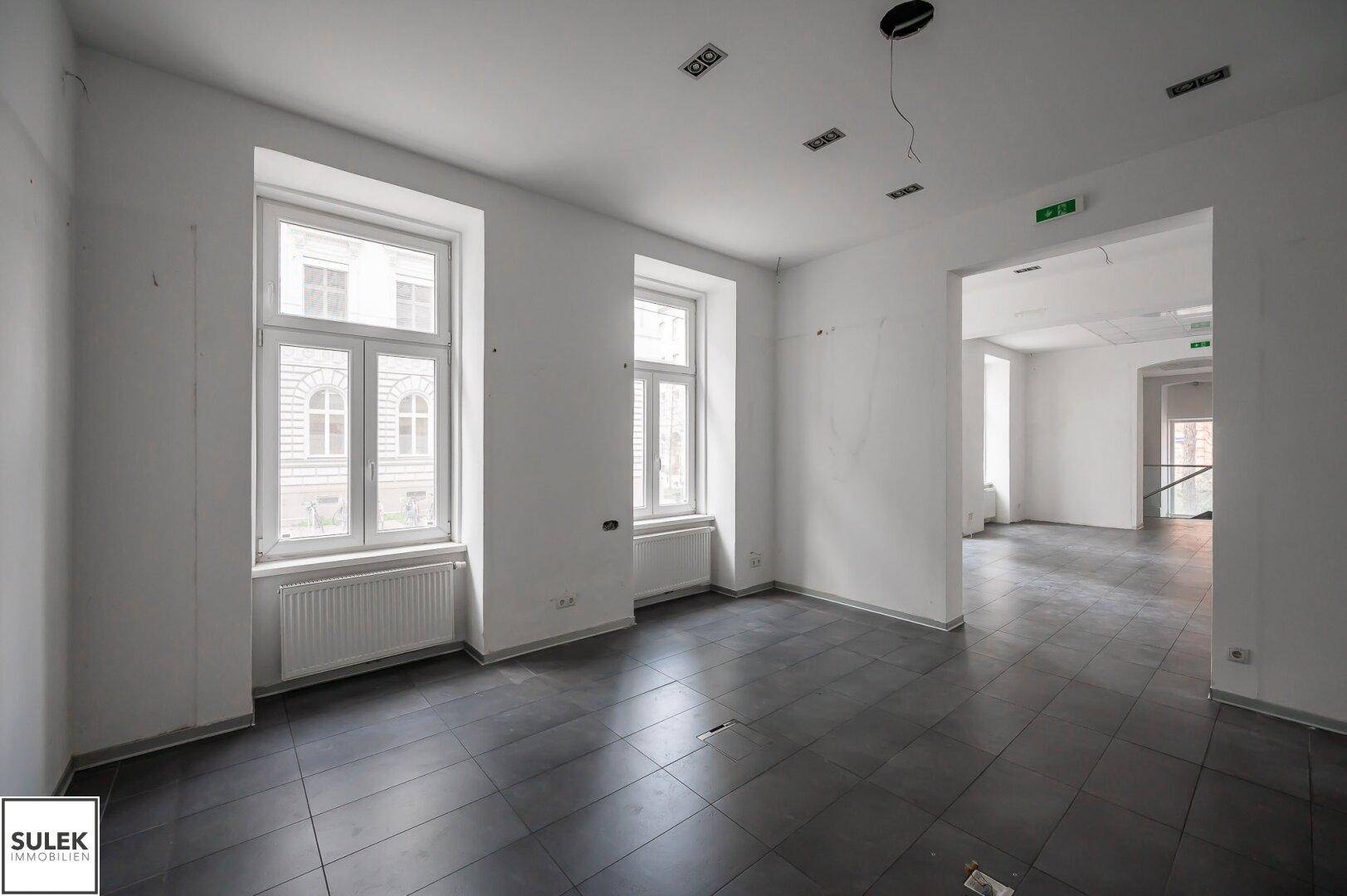 Heller Büroraum mit großen Fenstern, dunklen Fliesen und einer abgehängten Decke, bereit zur Renovierung.
