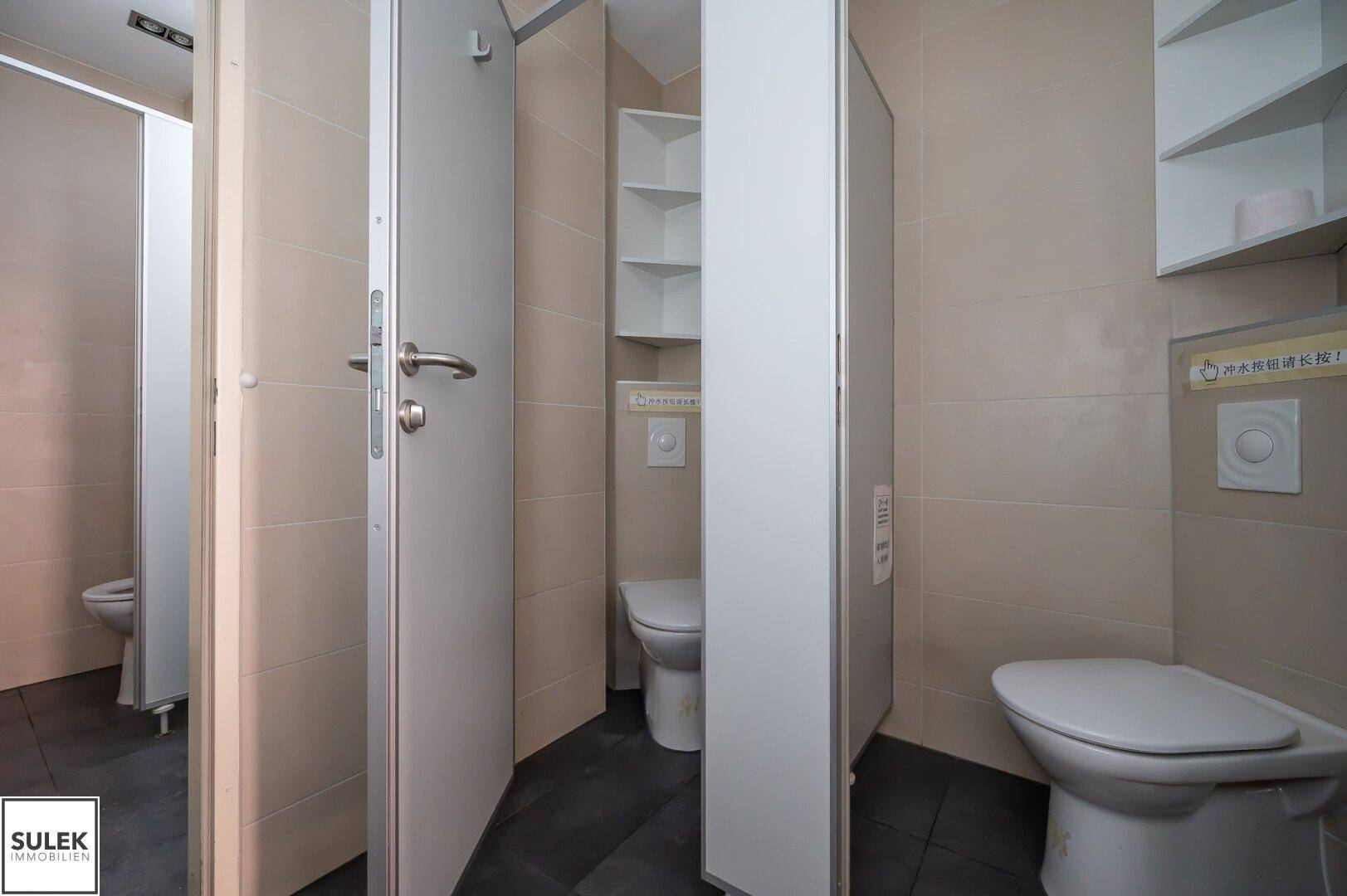 Mehrere WC-Kabinen in einem modernen Badezimmer mit beigefarbenen Fliesen und dunklem Boden.