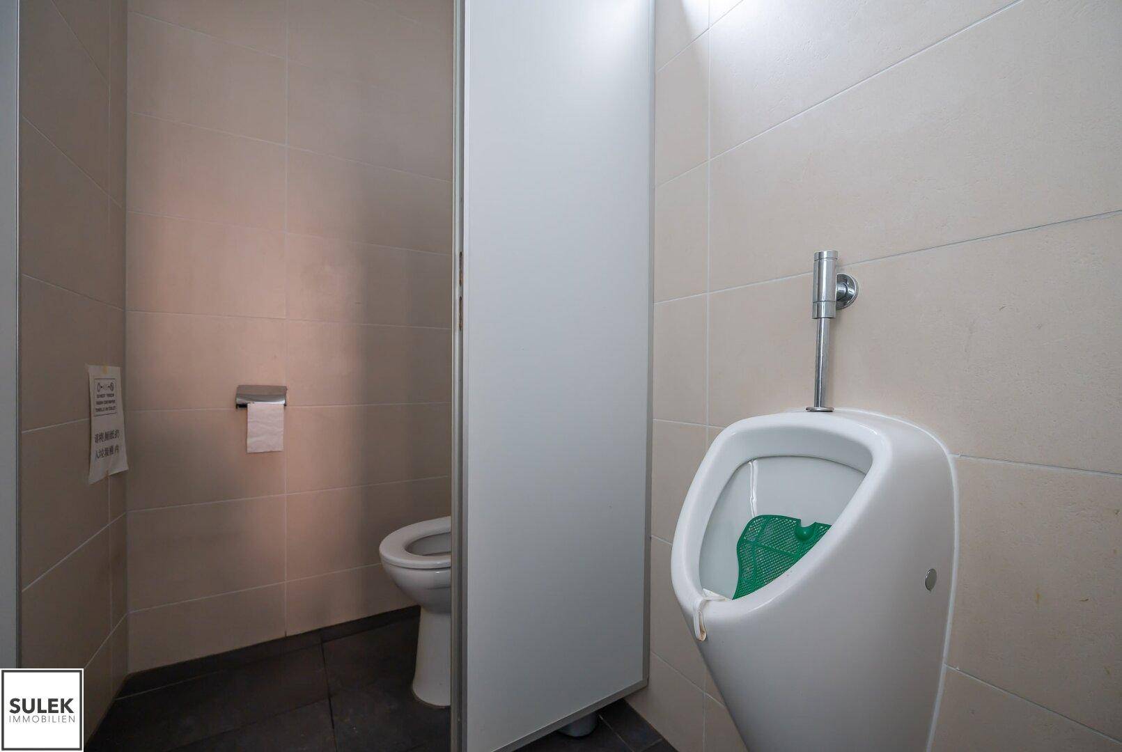 Moderne Herrentoilette mit Urinal und WC-Kabine, gefliesten Wänden und dunklem Boden.