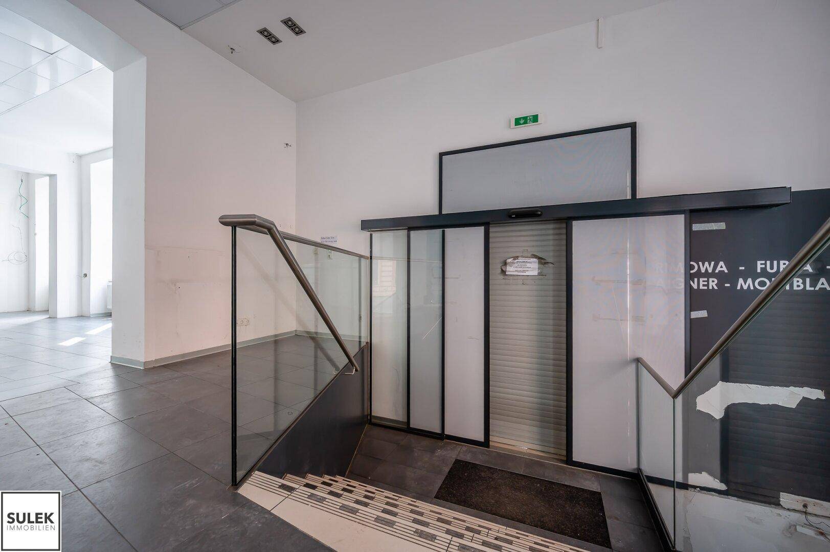 Eingangsbereich mit Treppe, Glasgeländer und dunklem Fliesenboden, der zu weiteren Etagen führt.